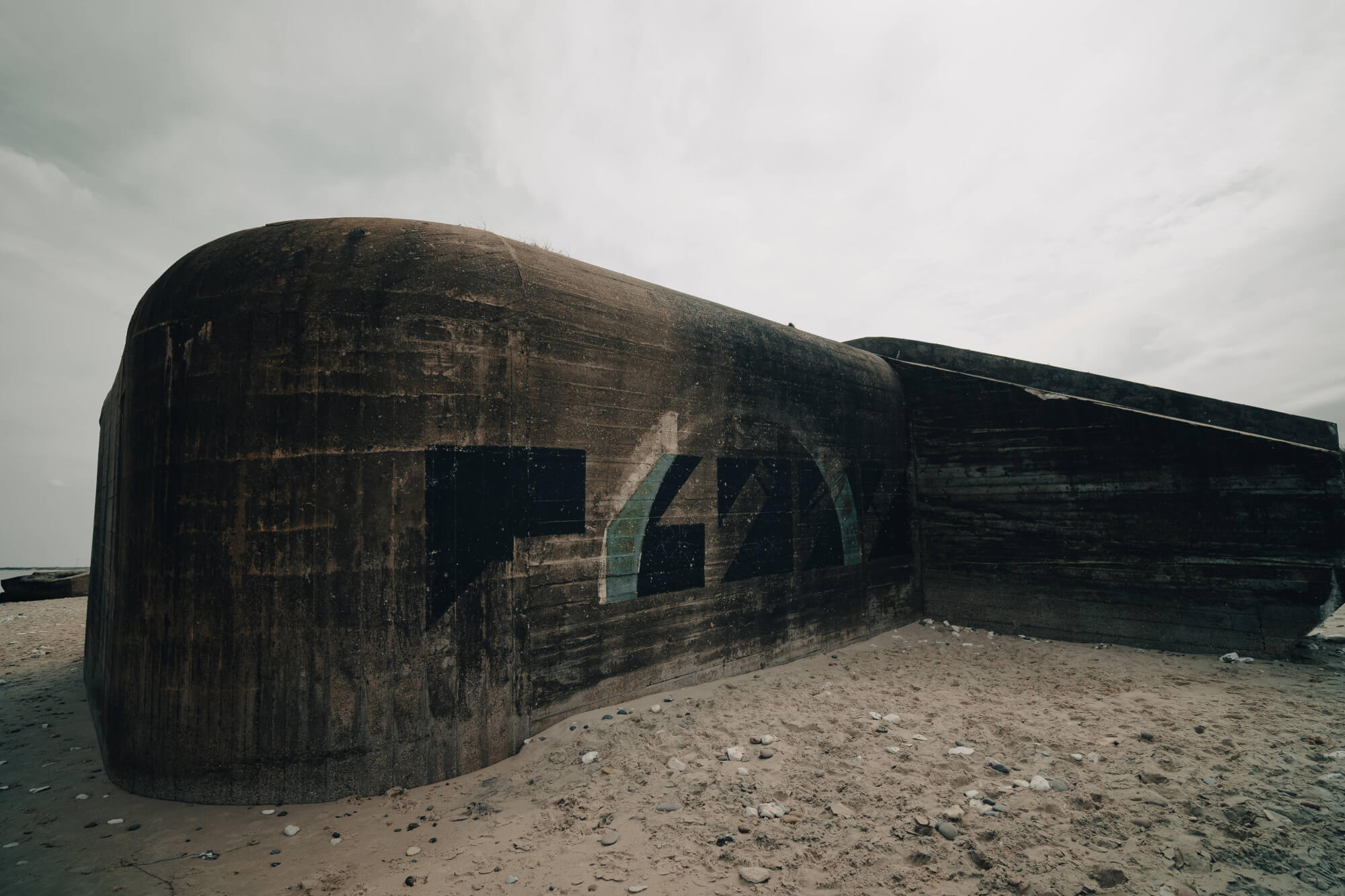 A large, weathered concrete bunker on a sandy beach under a cloudy sky, with faint graffiti visible on its surface.