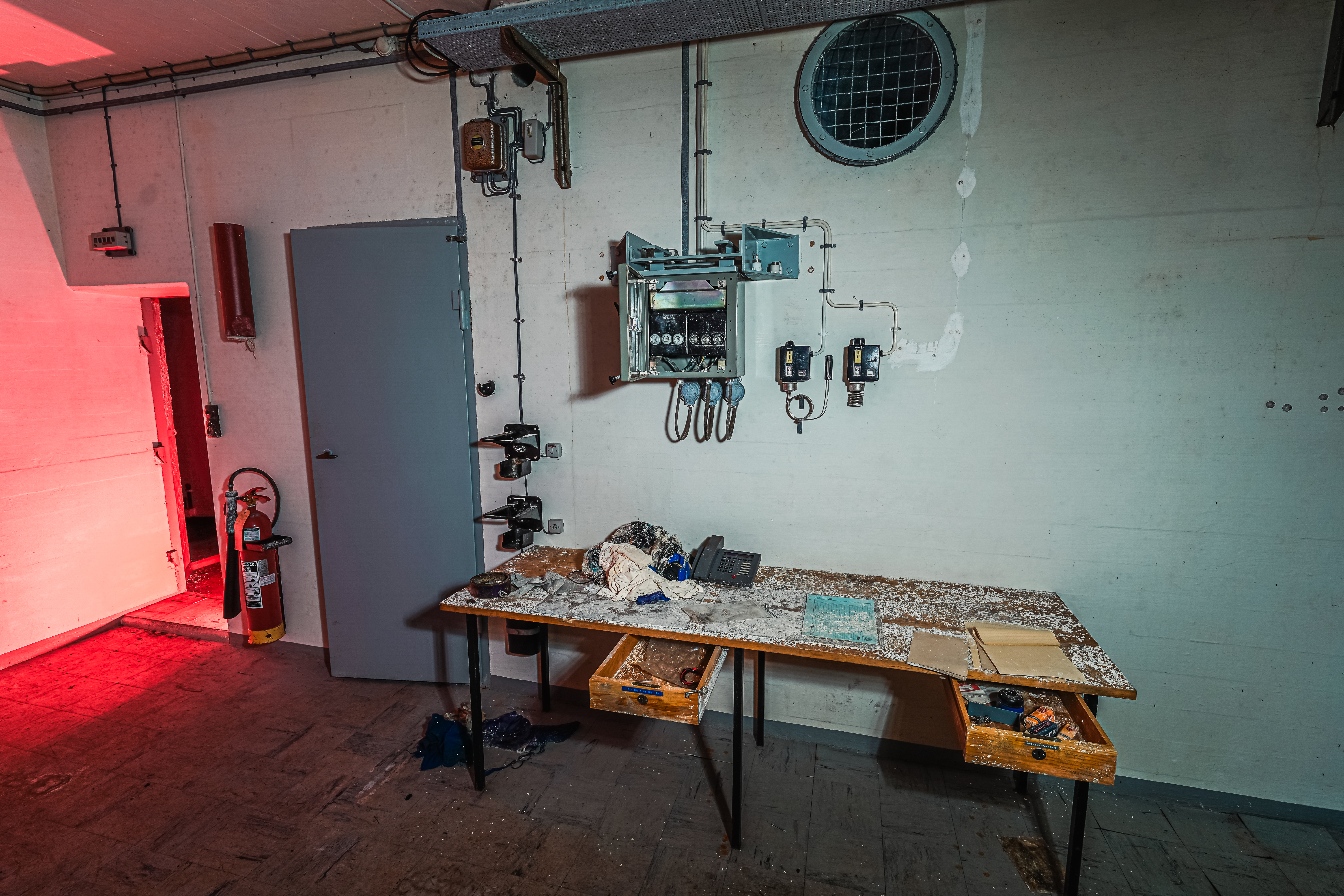 An industrial utility room with a wooden table covered in dust and clutter, an open control panel on the wall, and a red-lit doorway on the left.