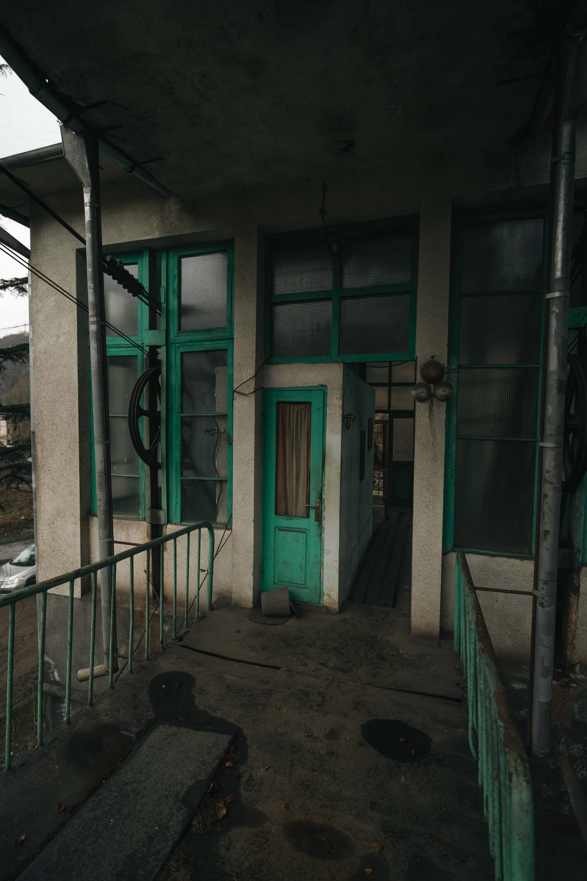 A weathered turquoise door leading into a slightly open doorway of a dilapidated building, surrounded by green-tinted windows and a worn railing. The ground is uneven, with patches of dark stains and scattered leaves, under an overcast sky.