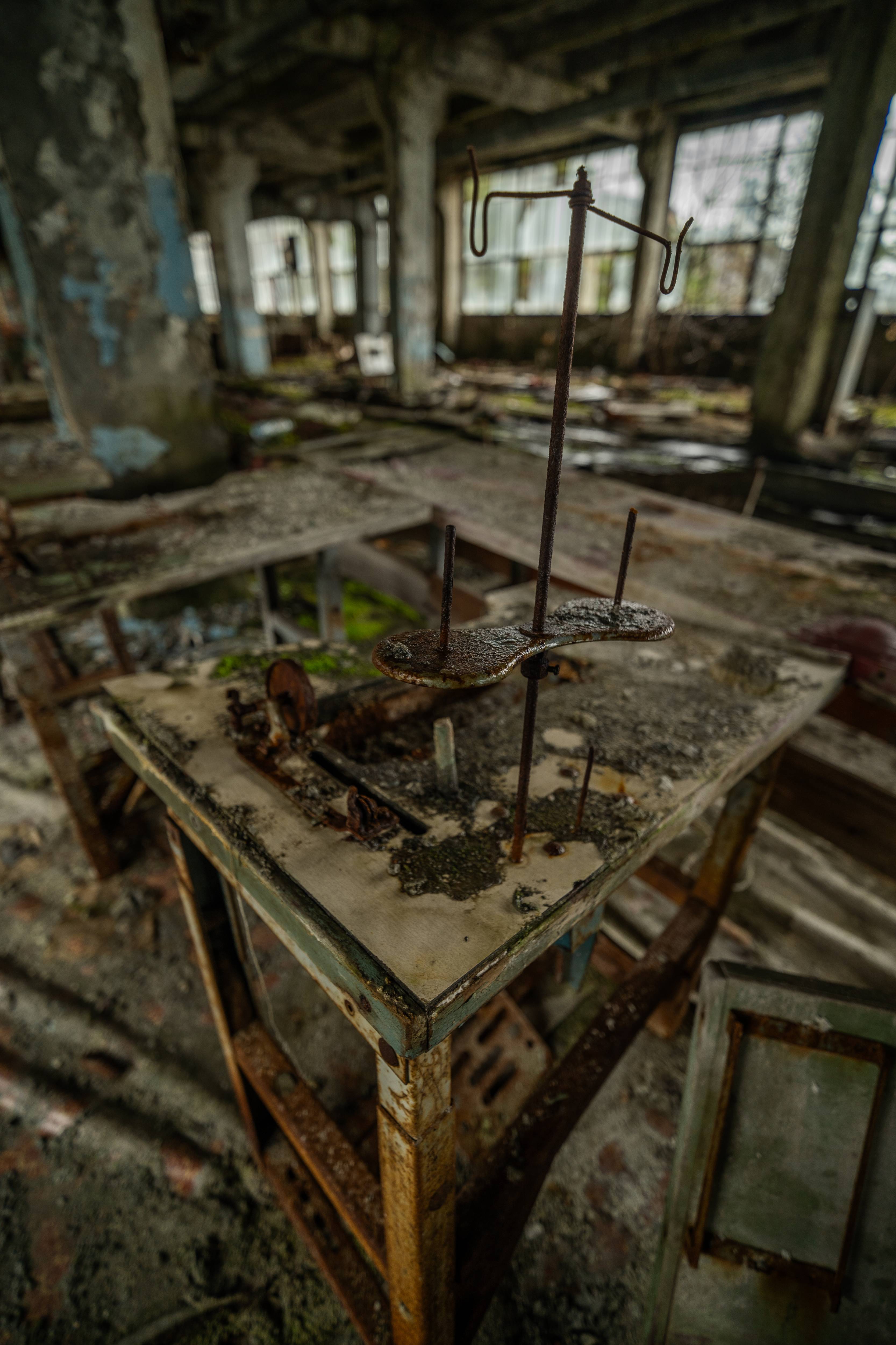 An abandoned industrial room with a focus on a rusty table and a support stand. The environment is dusty and overgrown, with large windows in the background letting in muted light.