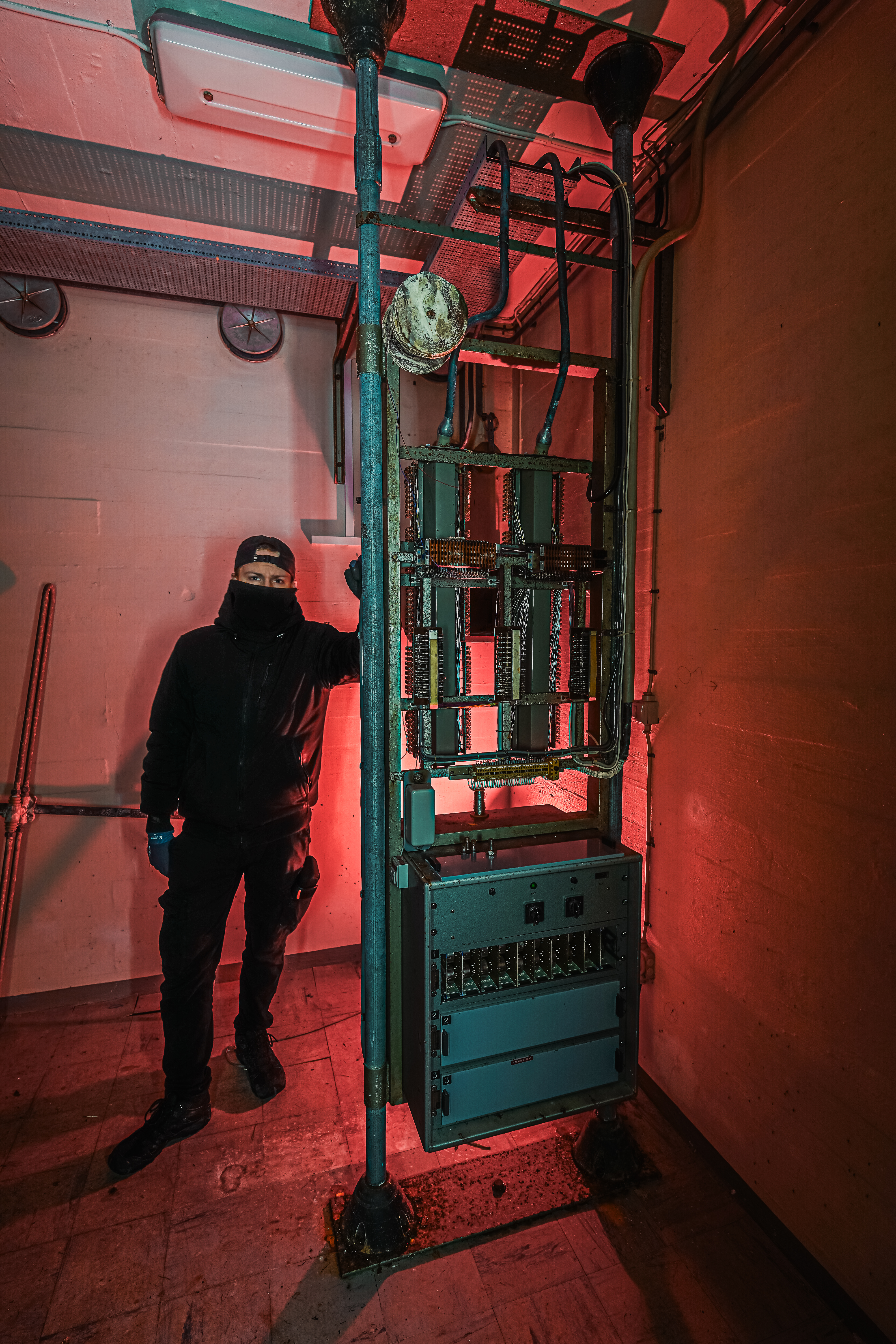 A person in a black outfit and mask stands beside an old electrical panel and pipes in a dimly lit room with red lighting.
