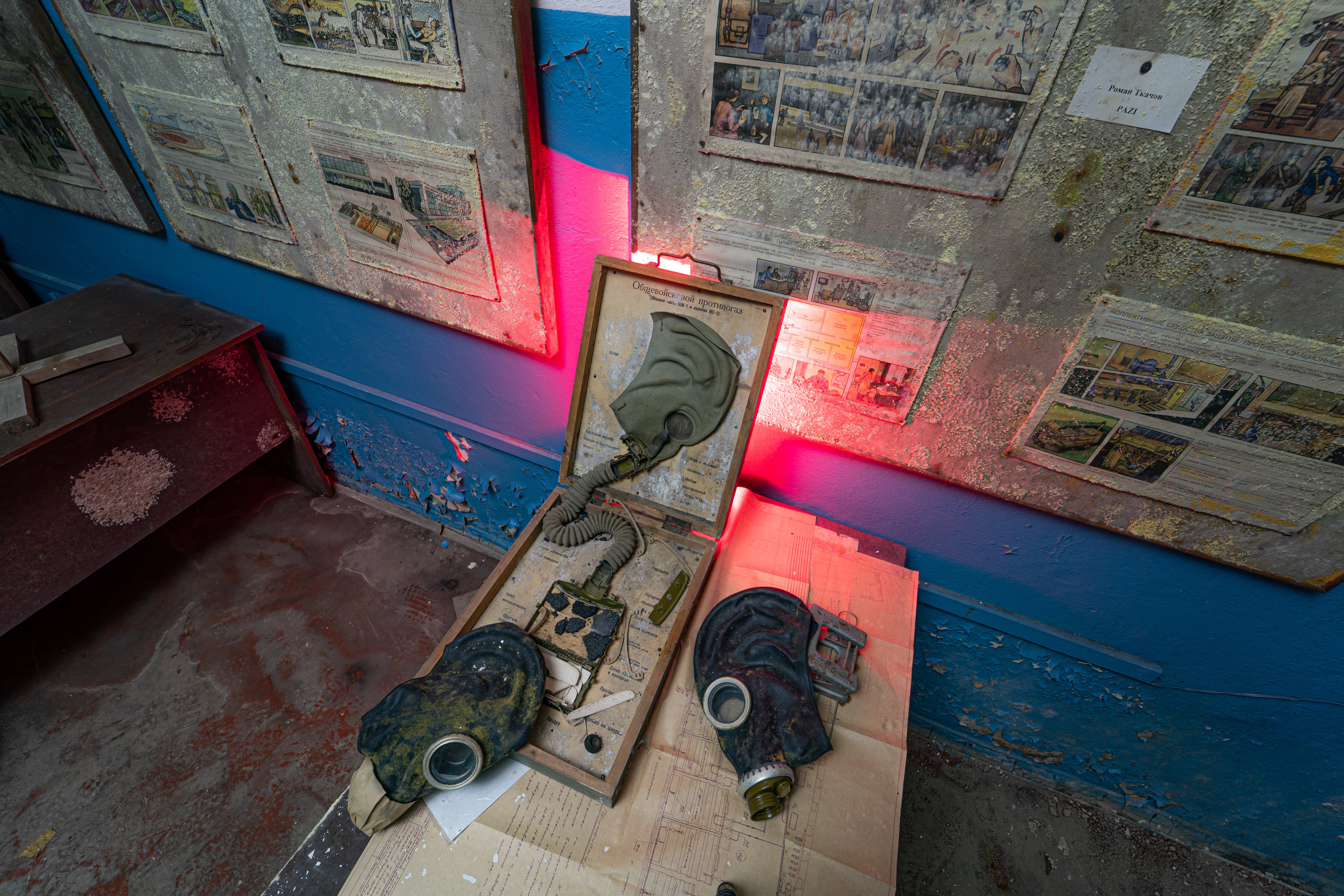 A collection of old gas masks displayed in an open case with red lighting, surrounded by dusty educational posters on a worn wall. The floor shows signs of neglect.