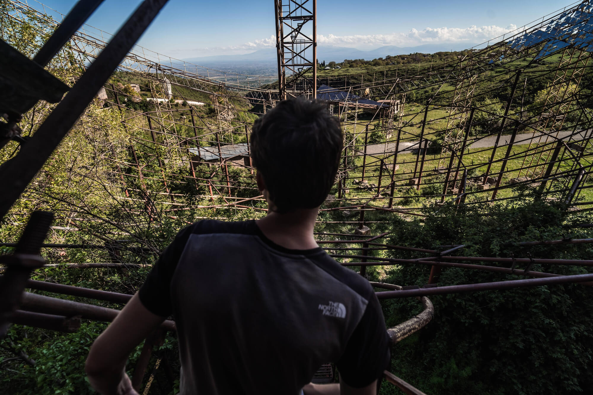 A person with short hair is facing away, standing near a dilapidated metal structure overgrown with plants, overlooking a vast green landscape under a clear sky.