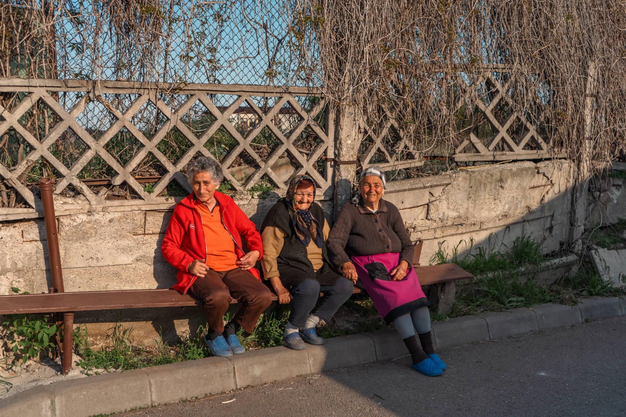 Three elderly women sitting on a wooden bench against a textured concrete wall with vines, wearing colorful traditional clothing. The scene is illuminated by warm sunlight, creating a cozy and friendly atmosphere.