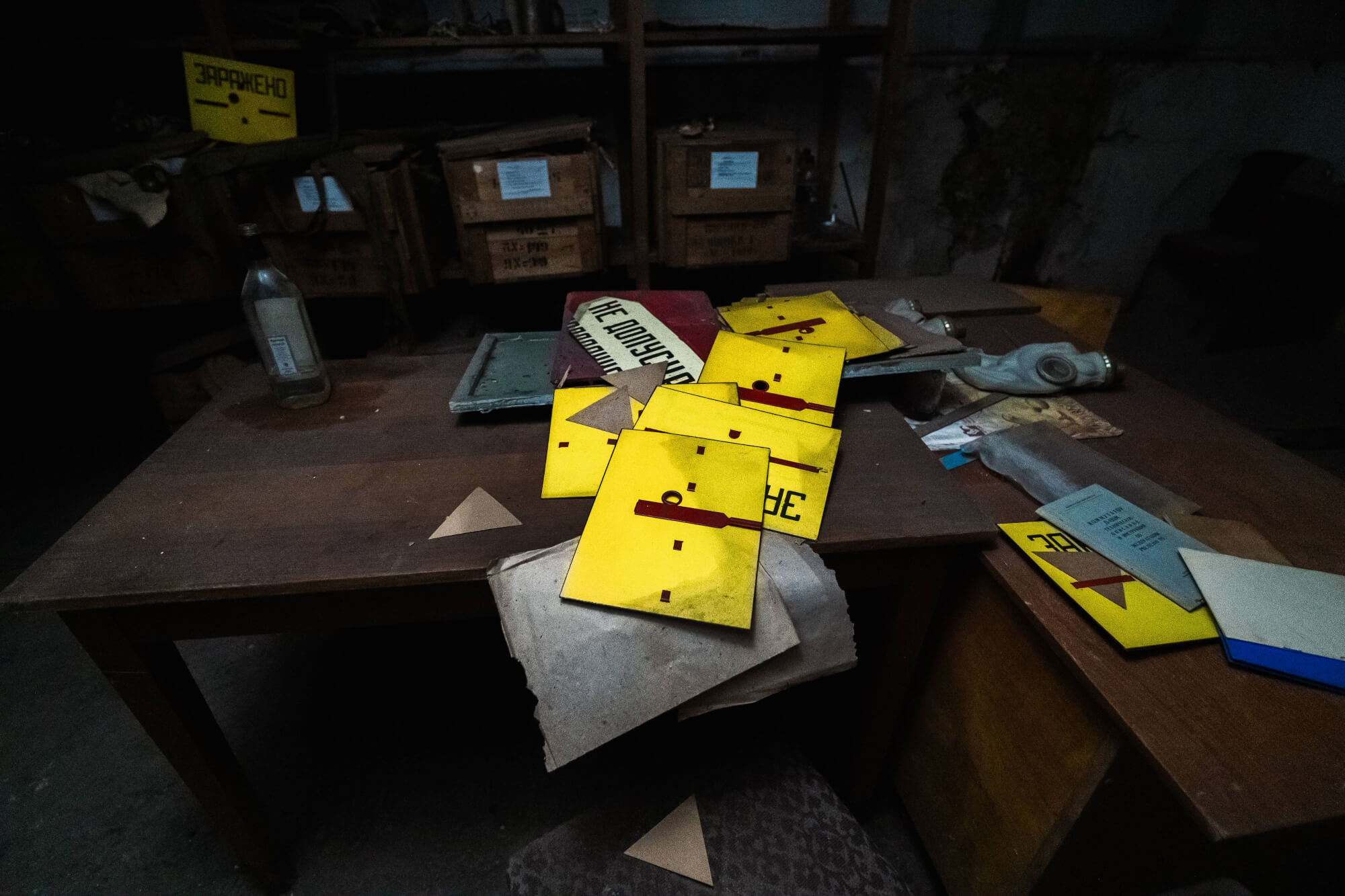 A wooden table cluttered with yellow warning signs, papers, and a gas mask in a dimly lit, abandoned room.