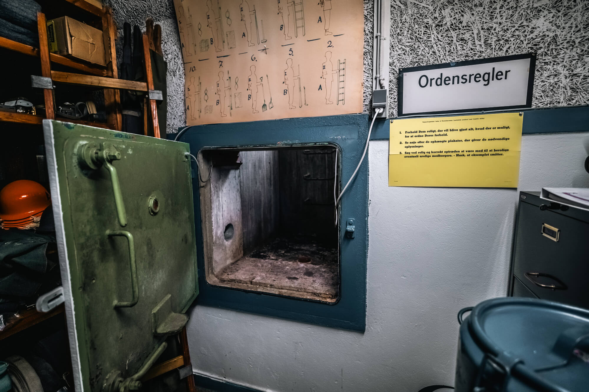 An open green metal door leading to a dark, empty space; behind it is a shelf with safety helmets and various tools, along with a sign and instructional diagrams on the wall.