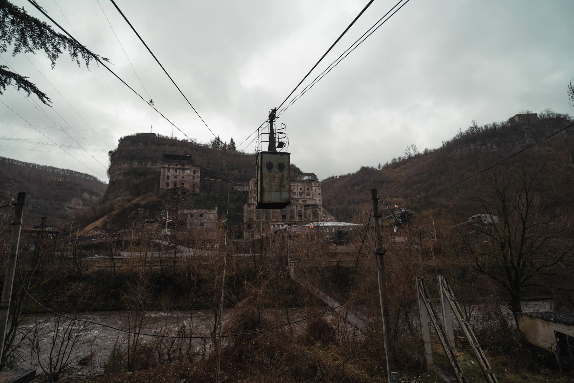 A rusted cable car hangs above a barren landscape featuring crumbling buildings on a hillside and a river below, under a cloudy sky.