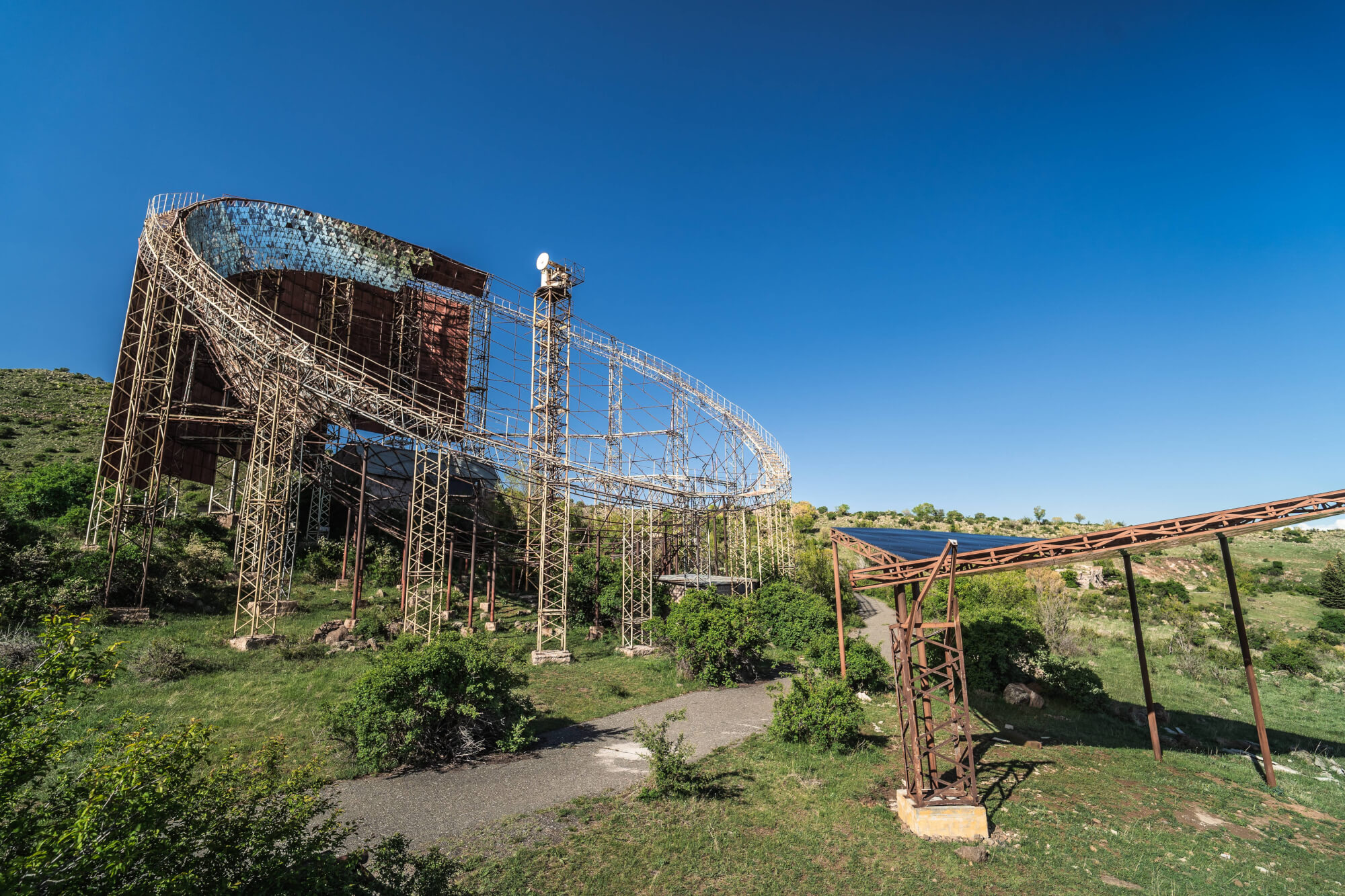 An abandoned metal structure with a curved design and rusted beams, surrounded by green bushes and a clear blue sky.