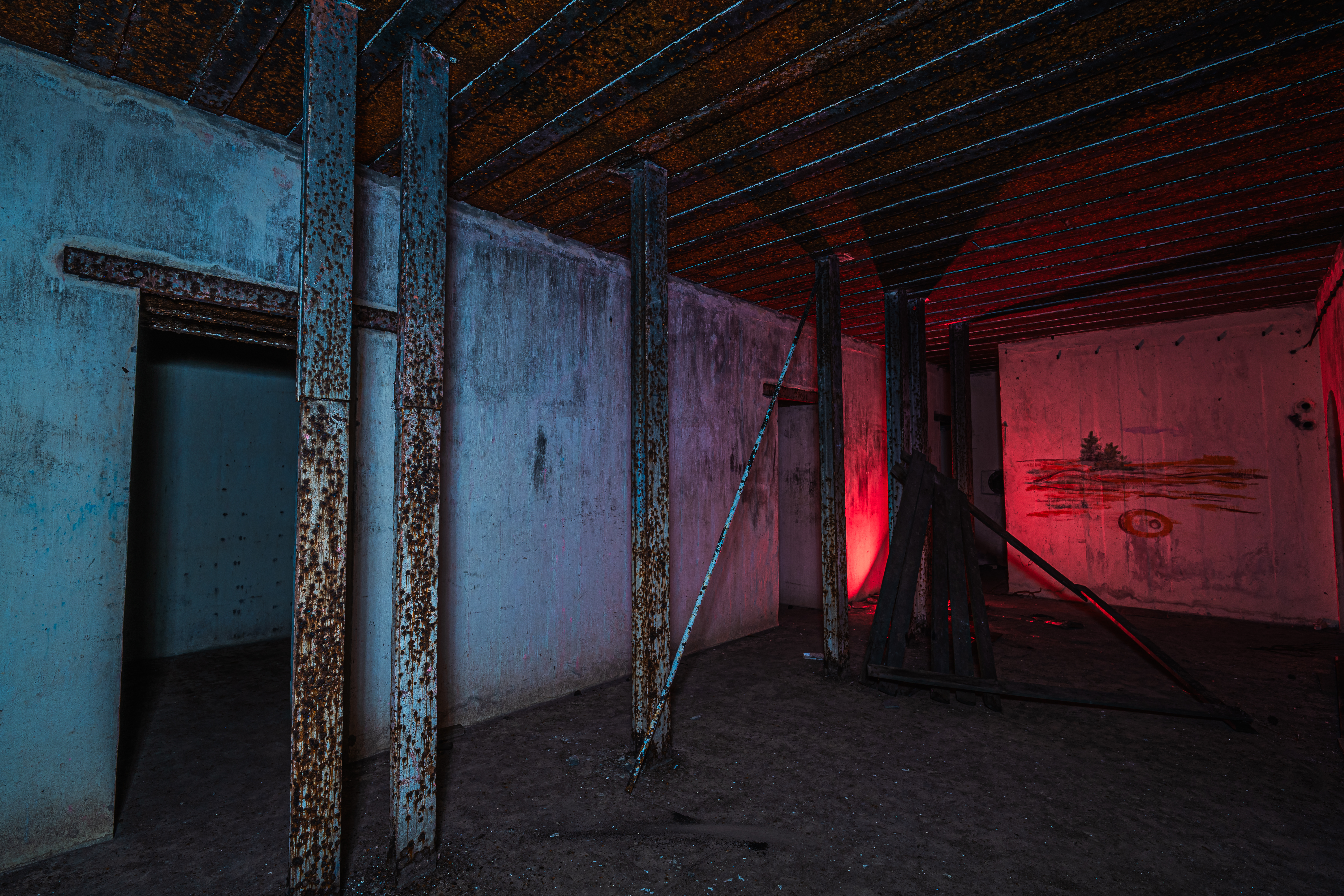 An abandoned room with rusting metal beams, peeling walls, and a faint red light illuminating one corner. The floor is dusty, and a wooden structure leans against a wall.