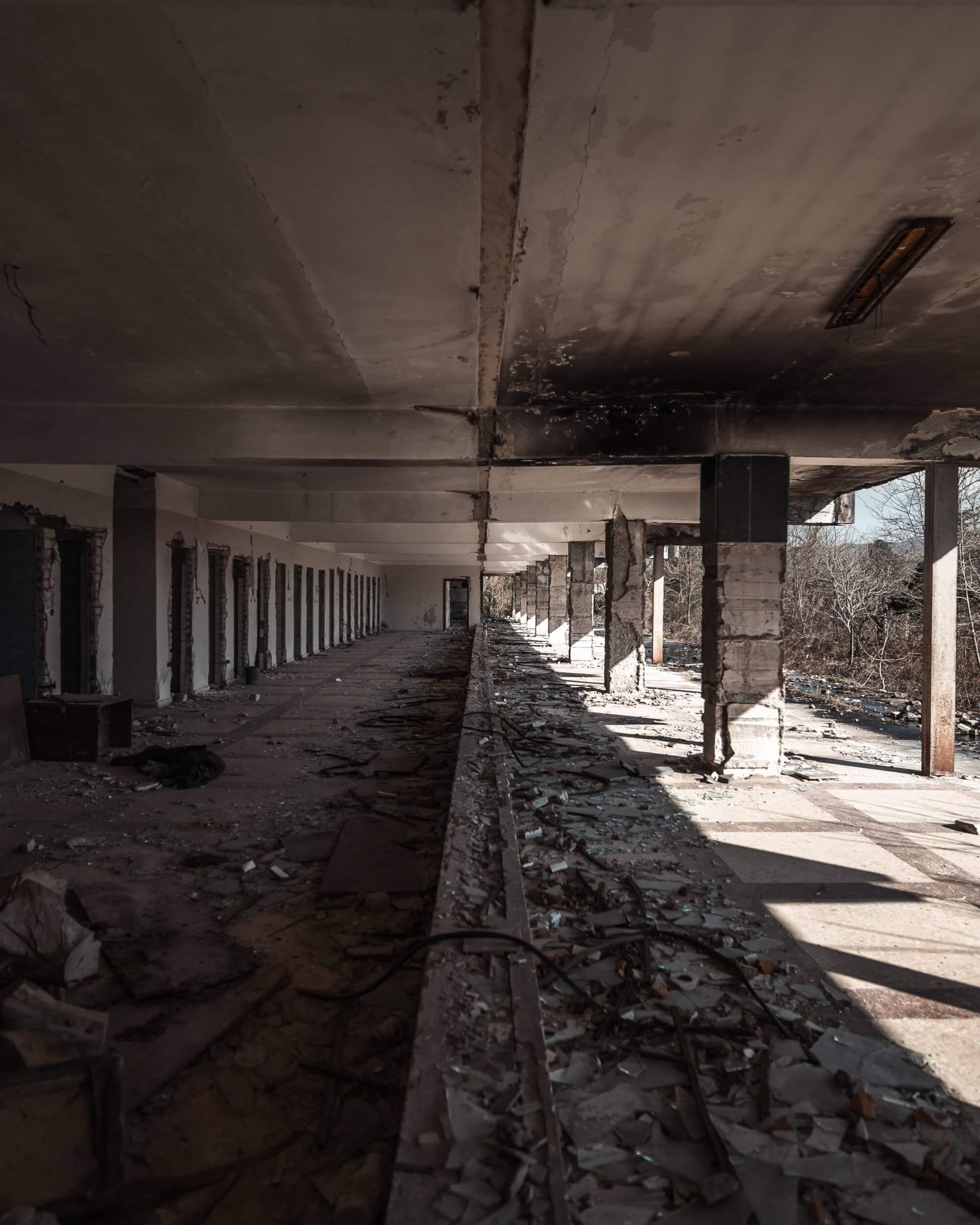 An empty, dilapidated corridor in a derelict building with broken windows, scattered debris, and shadowy light creating a somber and eerie atmosphere.