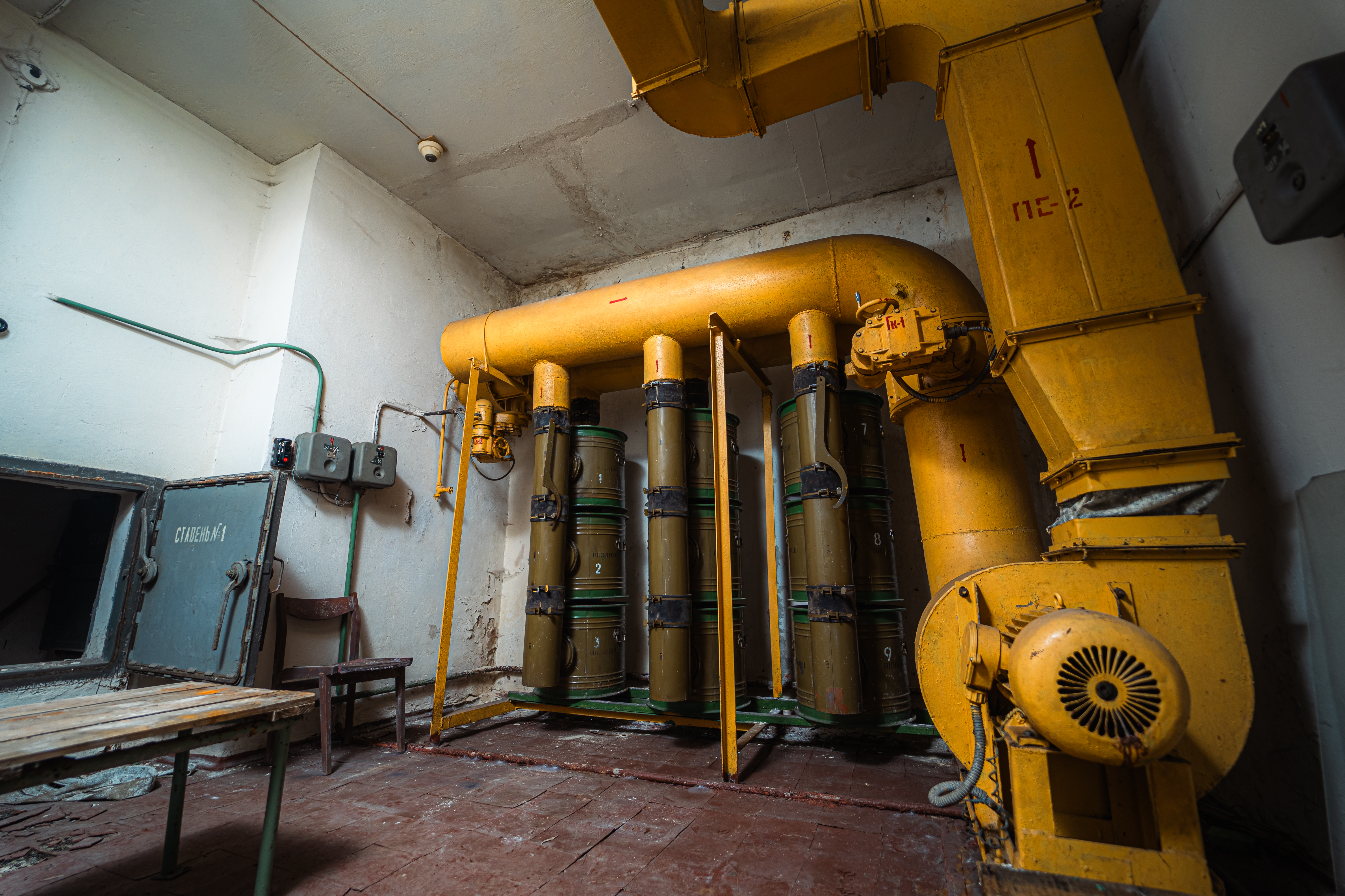 Interior view of an industrial room with yellow pipes, green barrels numbered from 1 to 9, a gray door, and a wooden chair and table.
