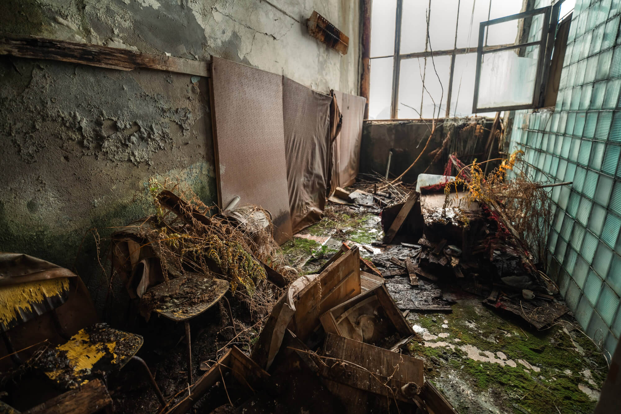 A cluttered and decaying room with disheveled furniture, vines, and a partially open large window letting in gentle light.