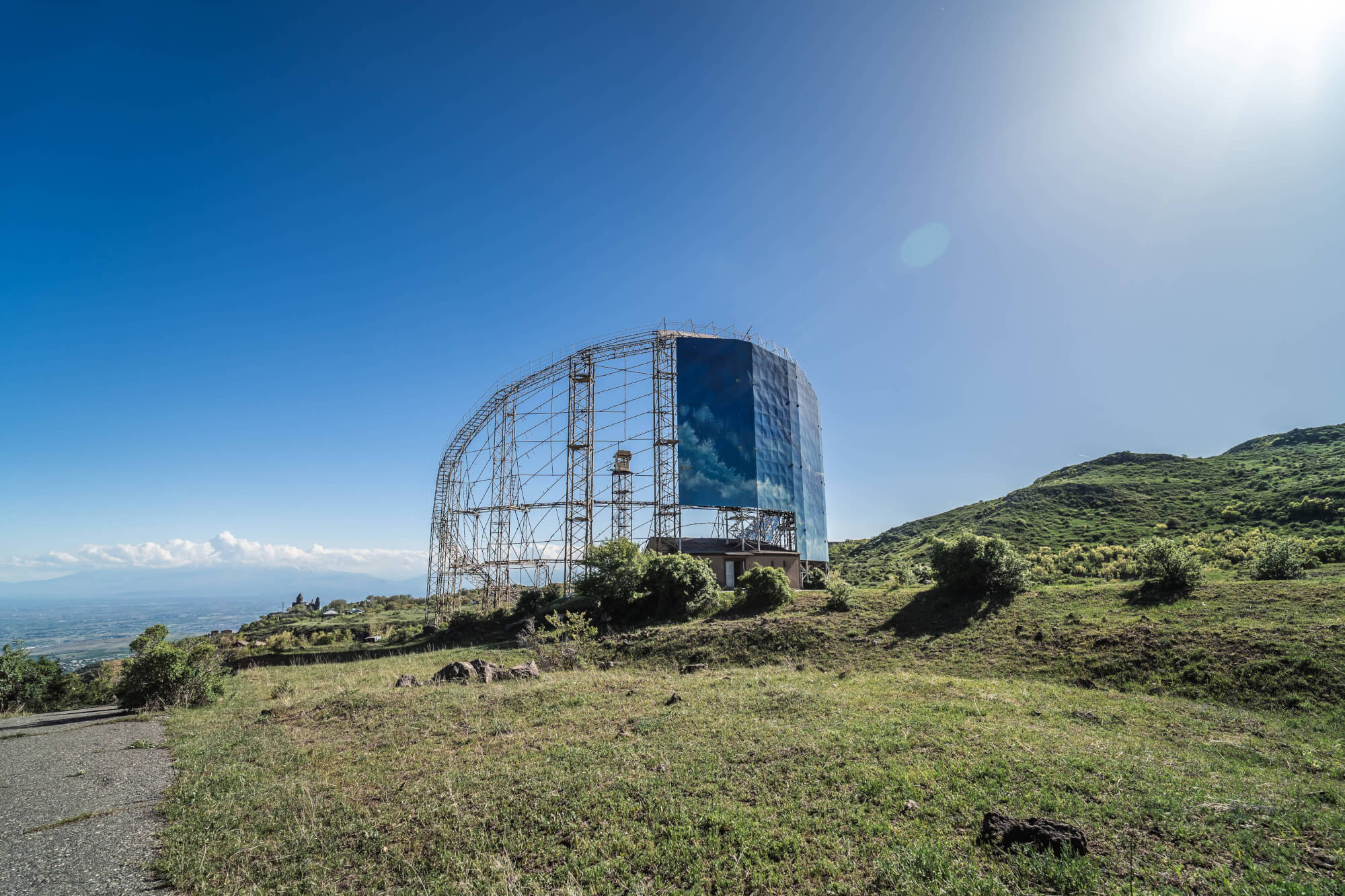 A large, partially constructed industrial structure against a clear blue sky, surrounded by green hills and grass.