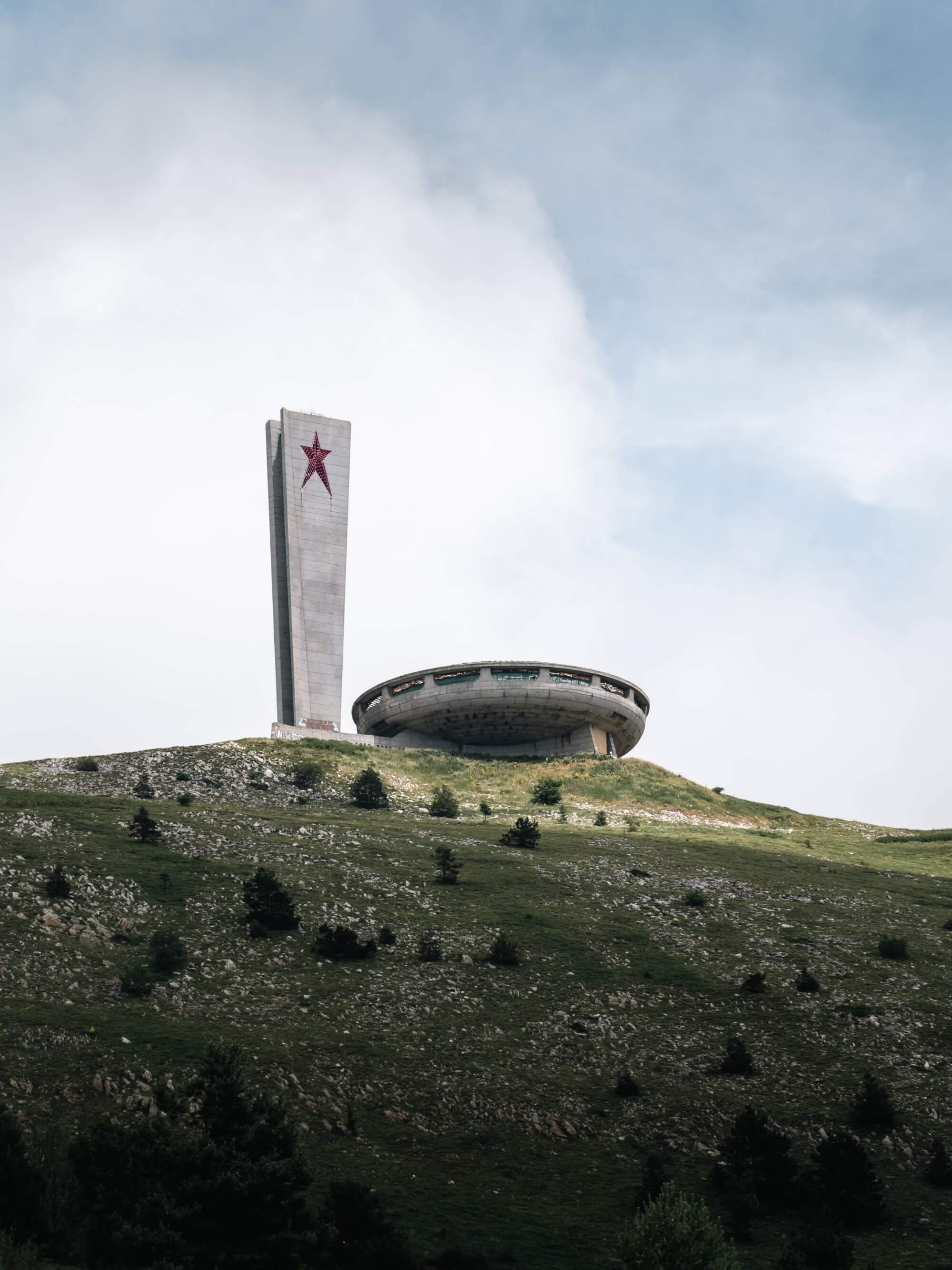 A tall concrete monument with a red star on top and a circular structure on a green hillside under a cloudy sky.