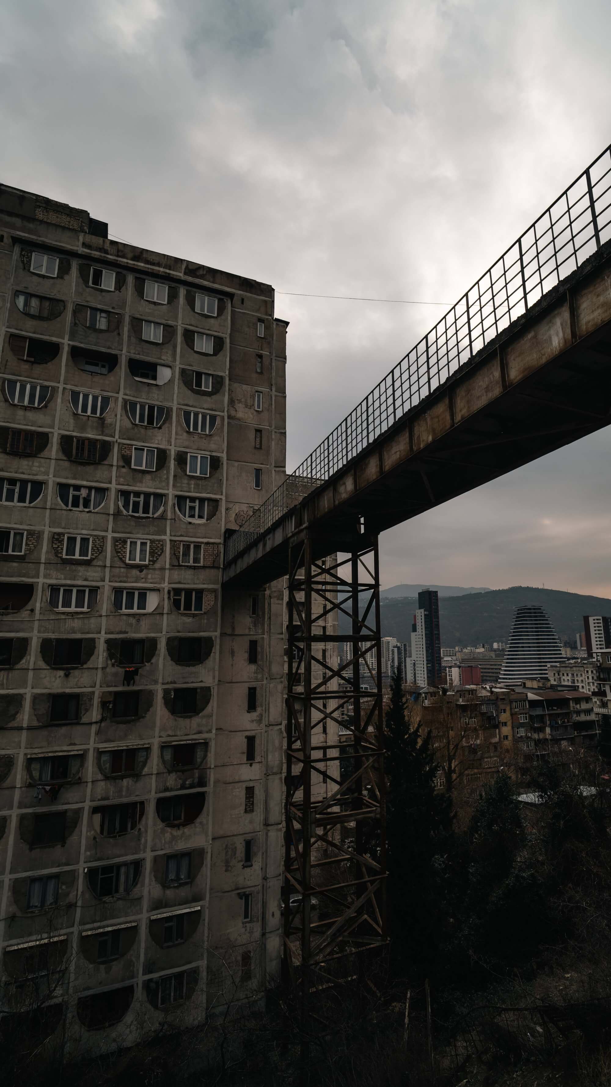 A dilapidated concrete apartment building adjacent to a rusted overpass, with modern skyscrapers in the background under a cloudy sky.