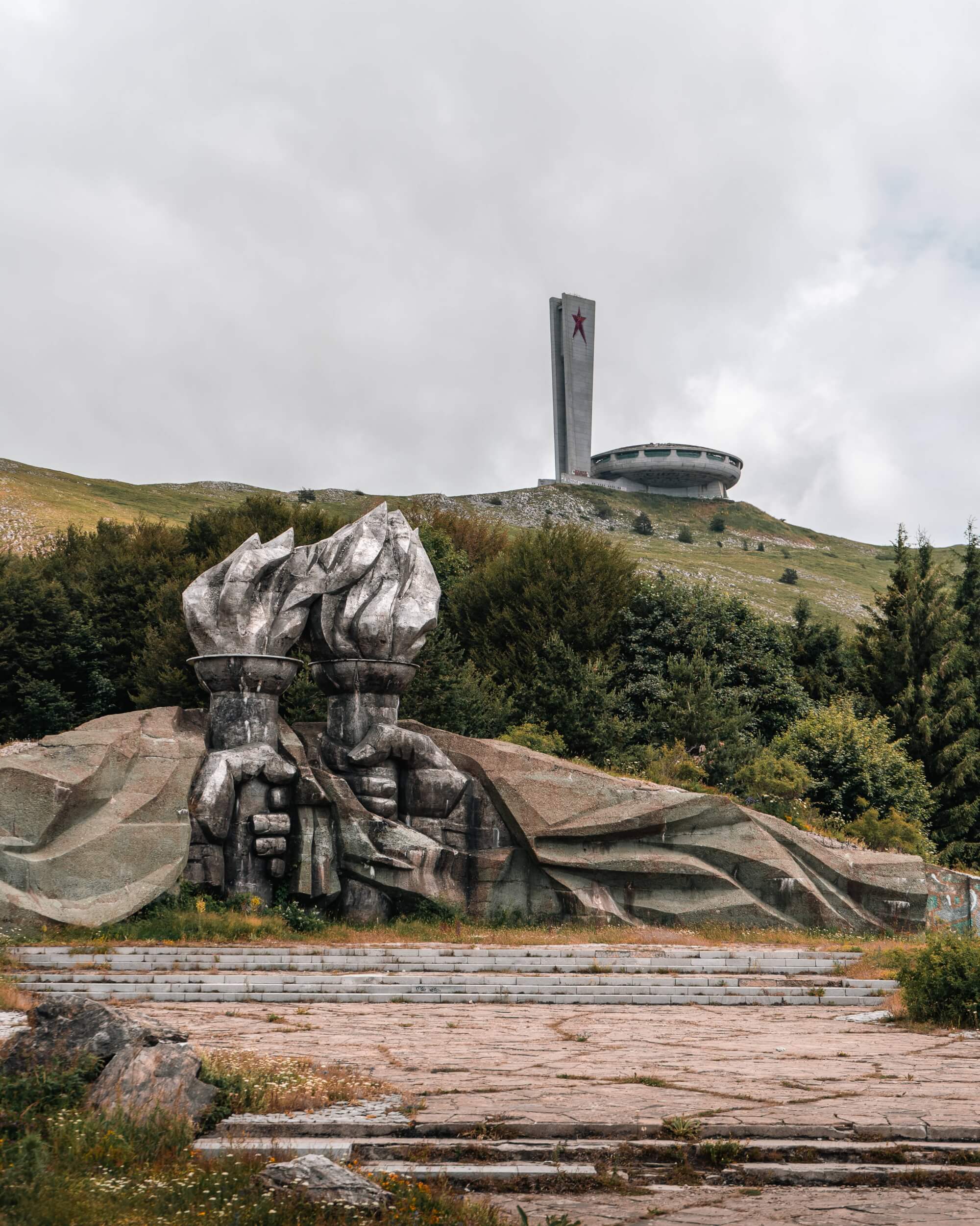 A large stone monument depicting two hands holding a torch in the foreground, with a modern building featuring a red star in the background, surrounded by green hills and cloudy skies.