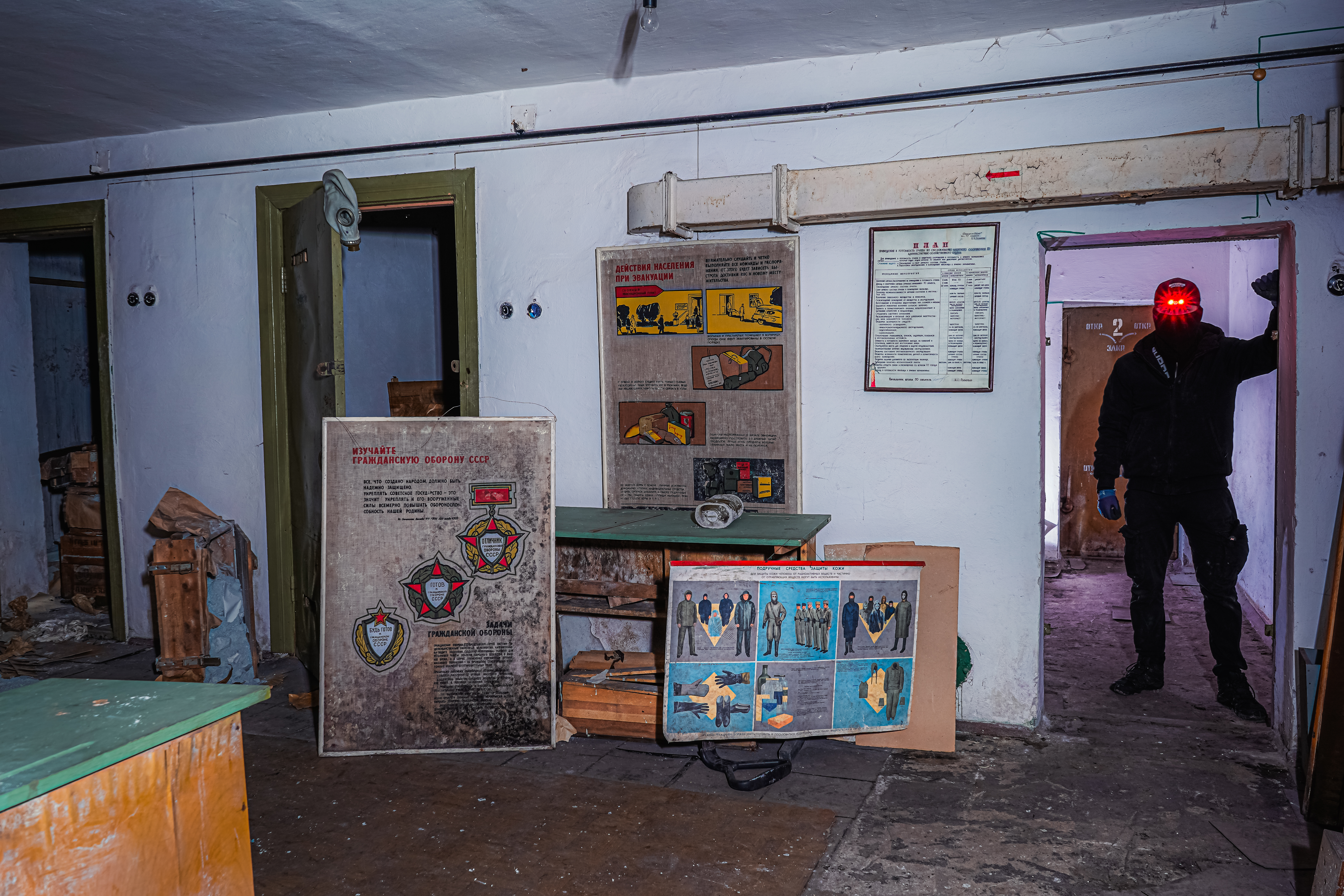 An abandoned room with peeling walls, scattered debris, and faded informational posters. A shadowy figure in dark clothing stands in a doorway, illuminated by a red light.