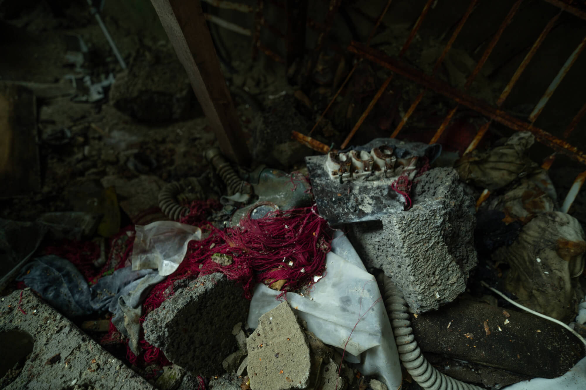 A cluttered scene featuring concrete blocks, tangled red and grey textiles, and discarded items scattered across a dark, shadowy space.