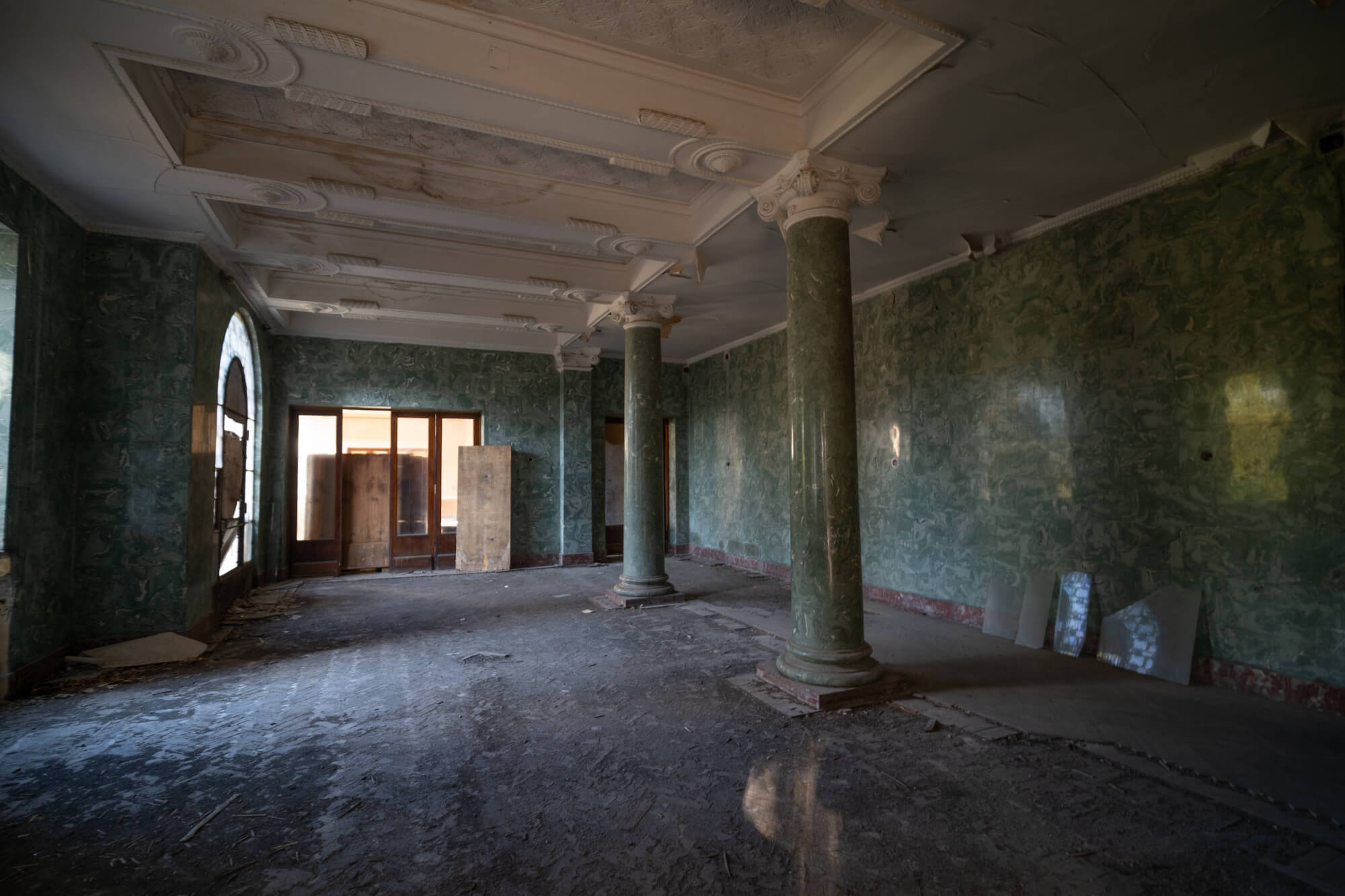 Interior of an abandoned room with green patterned walls, marble columns, wooden doors, and scattered debris on the floor.
