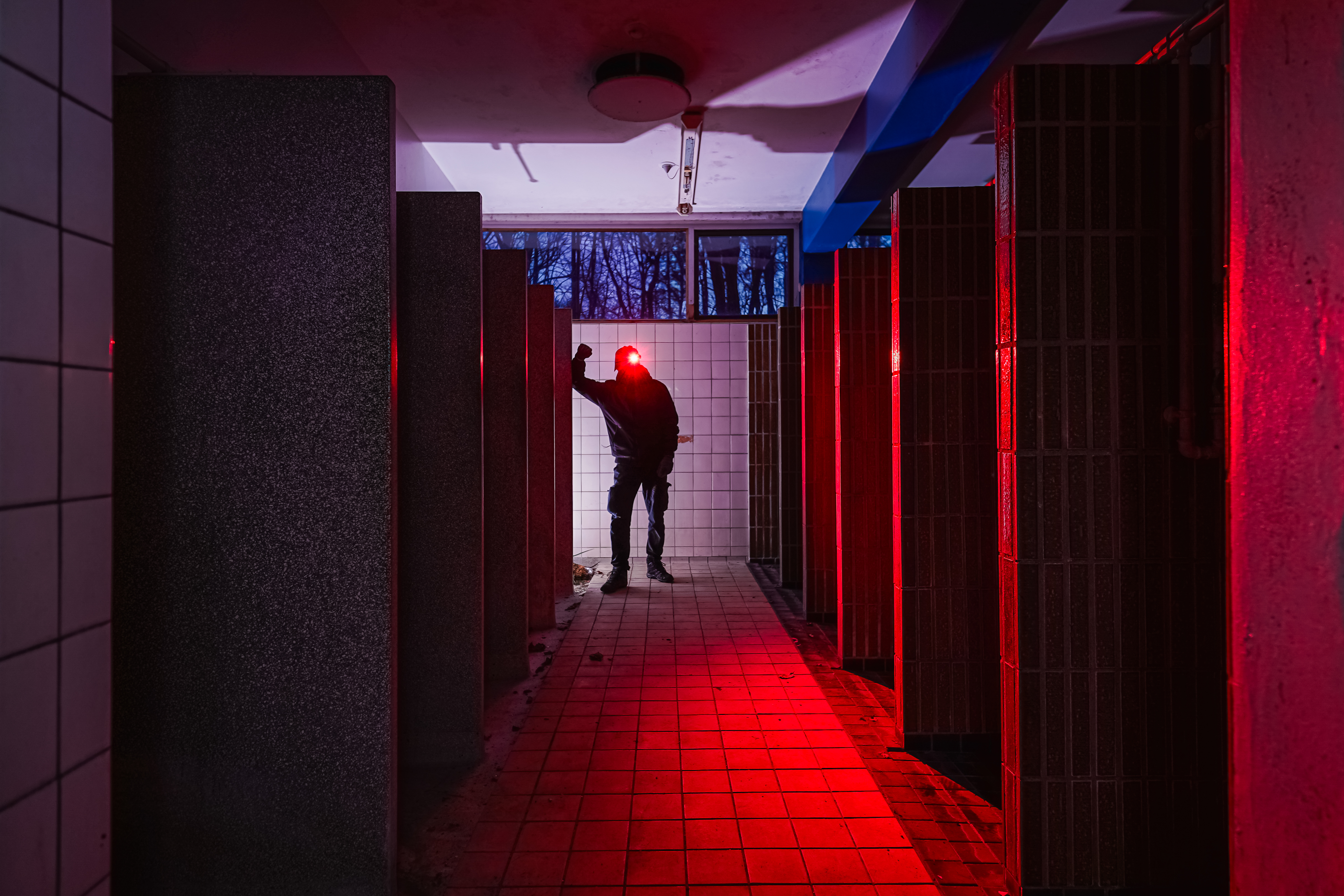 A dimly lit restroom with stone partitions, featuring a silhouetted figure against white tiles illuminated by red light.