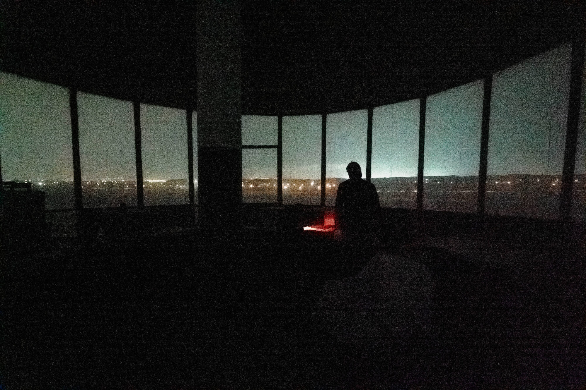 A dark room with large windows showing a cityscape at night; a silhouette of a person stands near a glowing red object.