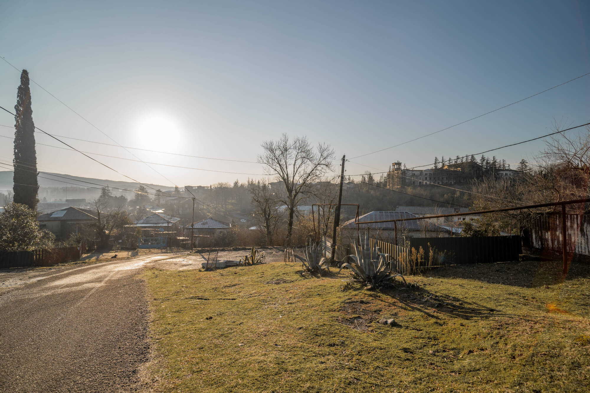 A gravel road winding through a green landscape with quiet houses and trees under a clear sky at sunrise.