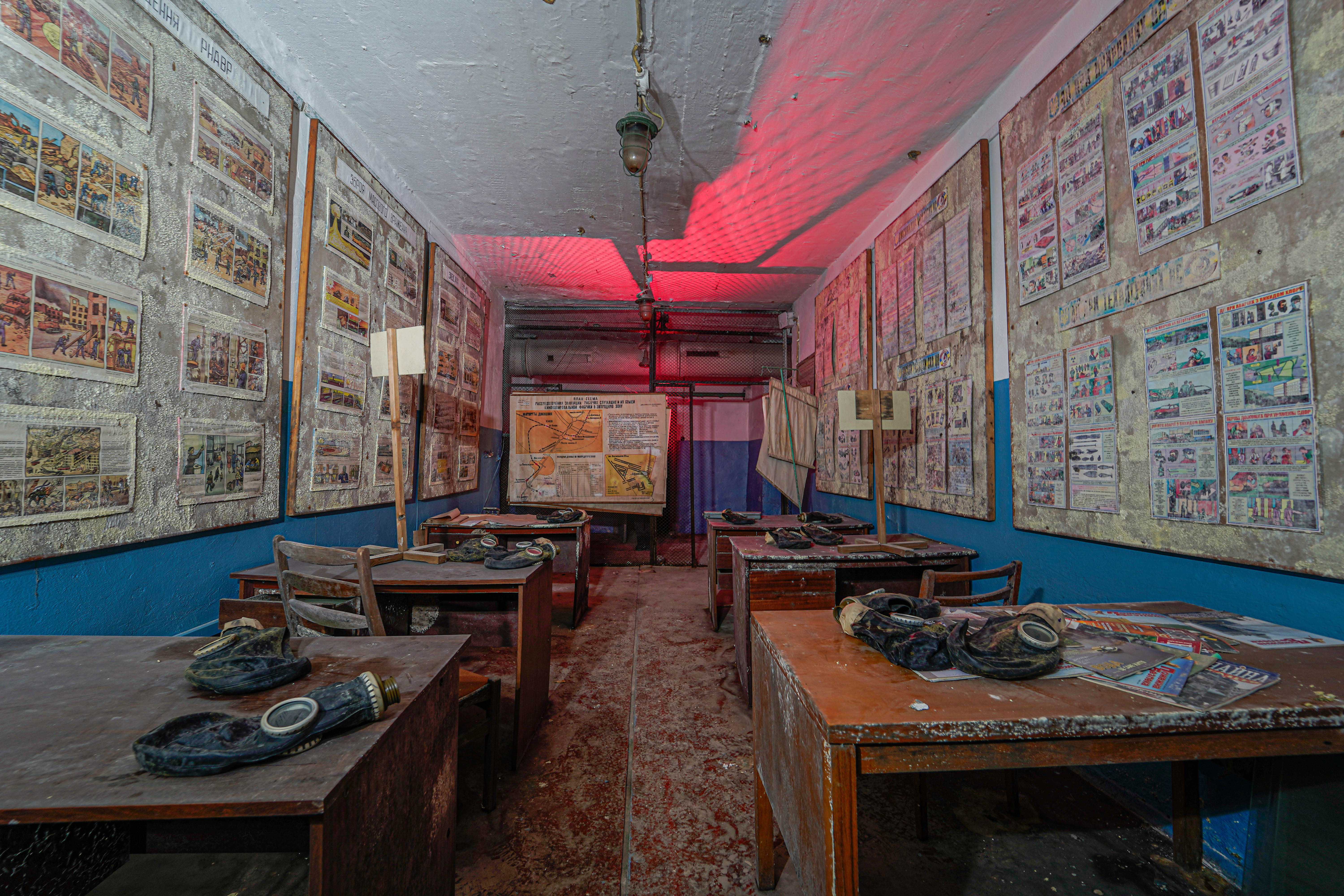 An abandoned classroom with worn wooden desks, gas masks on the tables, and walls filled with old posters and illustrations. A red light illuminates the space, enhancing the atmosphere of neglect.