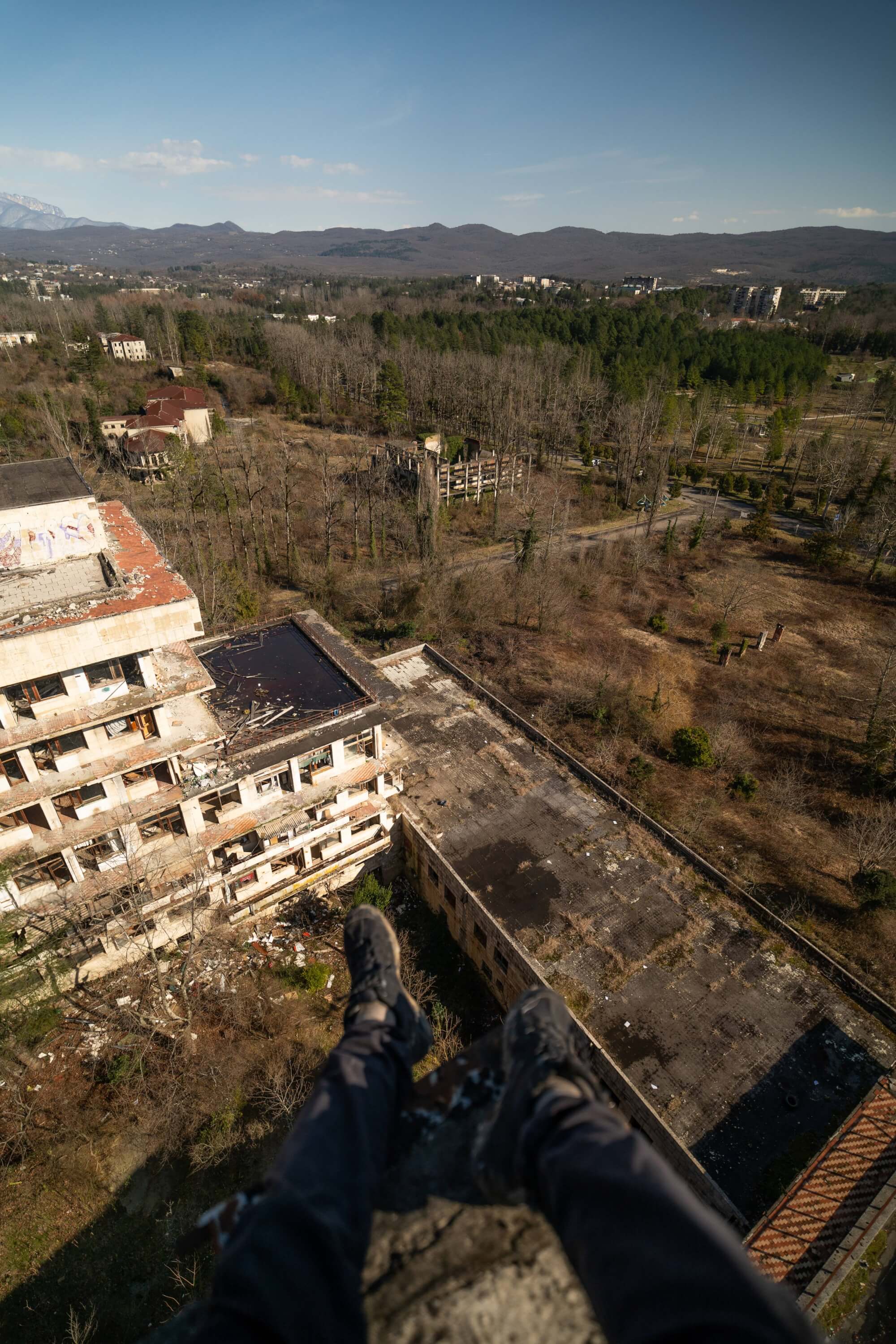 A pair of feet in black sneakers hanging over the edge of a dilapidated building, with an expansive view of overgrown land and abandoned structures below, under a clear blue sky.
