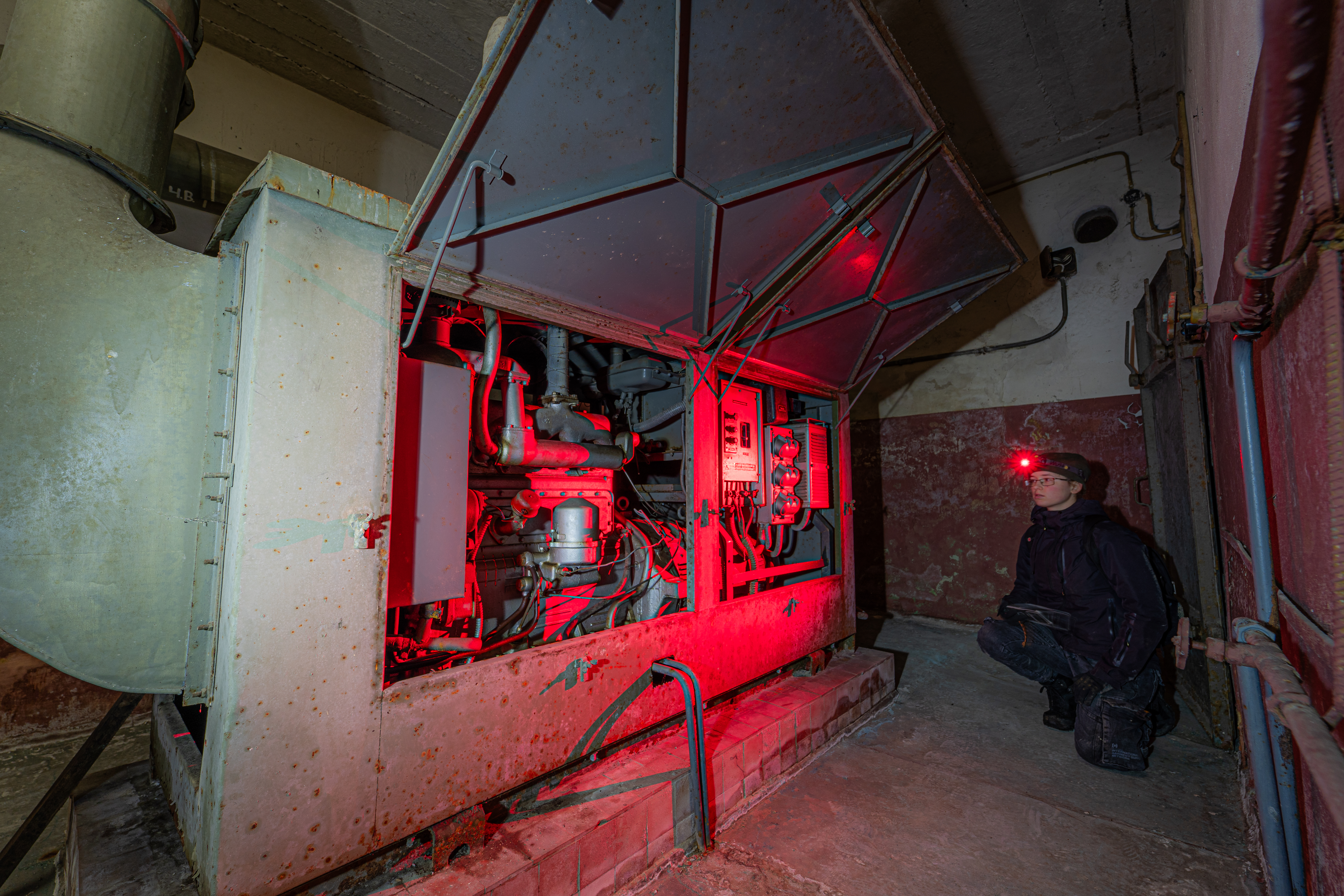 An old industrial generator surrounded by red lighting, with a person kneeling next to it. The generator is rusted, and the space has a worn-out appearance. The person is wearing a headlamp, suggesting exploration.