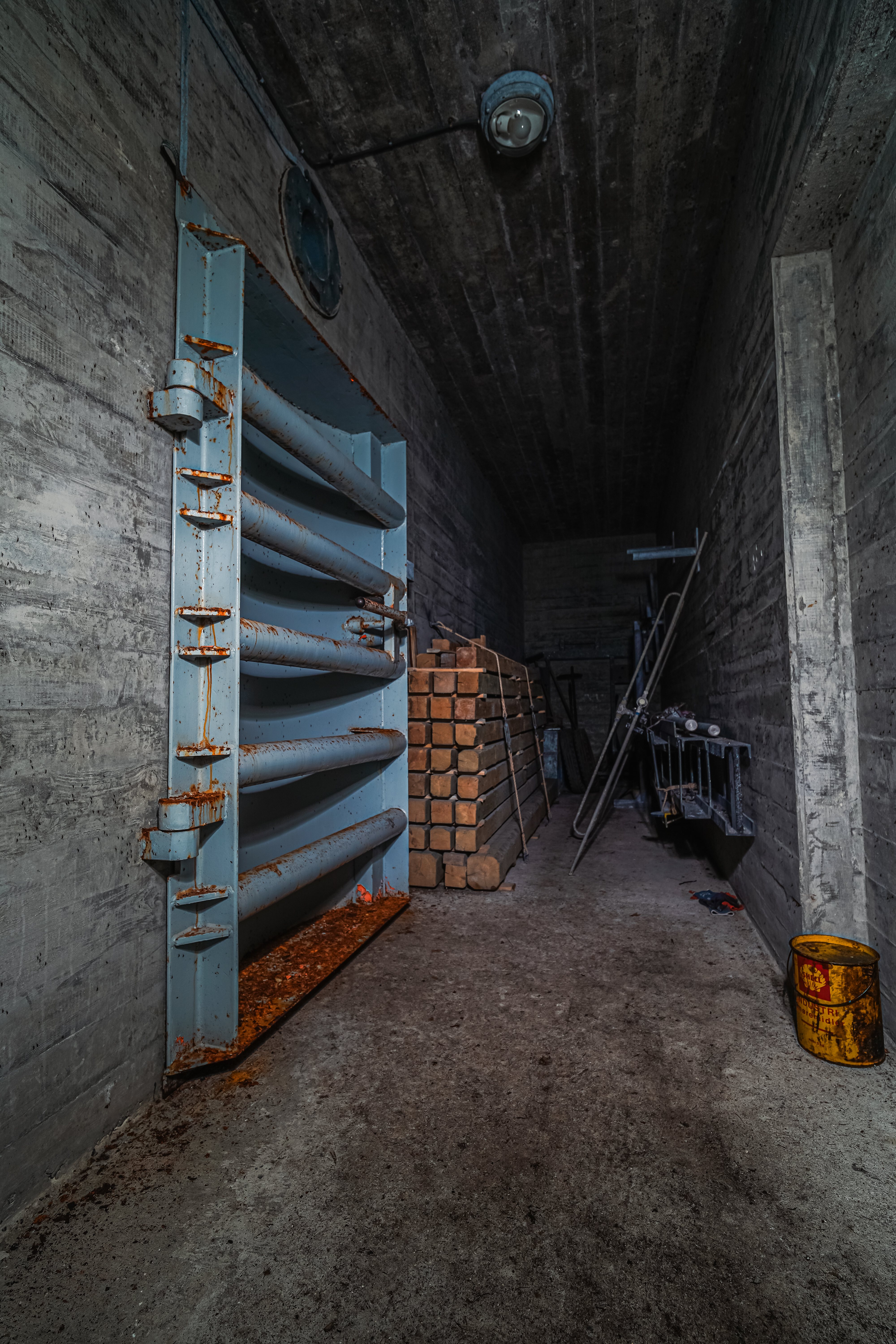 Interior of a dimly lit concrete hallway featuring a rusted blue steel door, stacks of wooden blocks, metal rods, and a yellow barrel on the floor, suggesting a once-utilized industrial space.