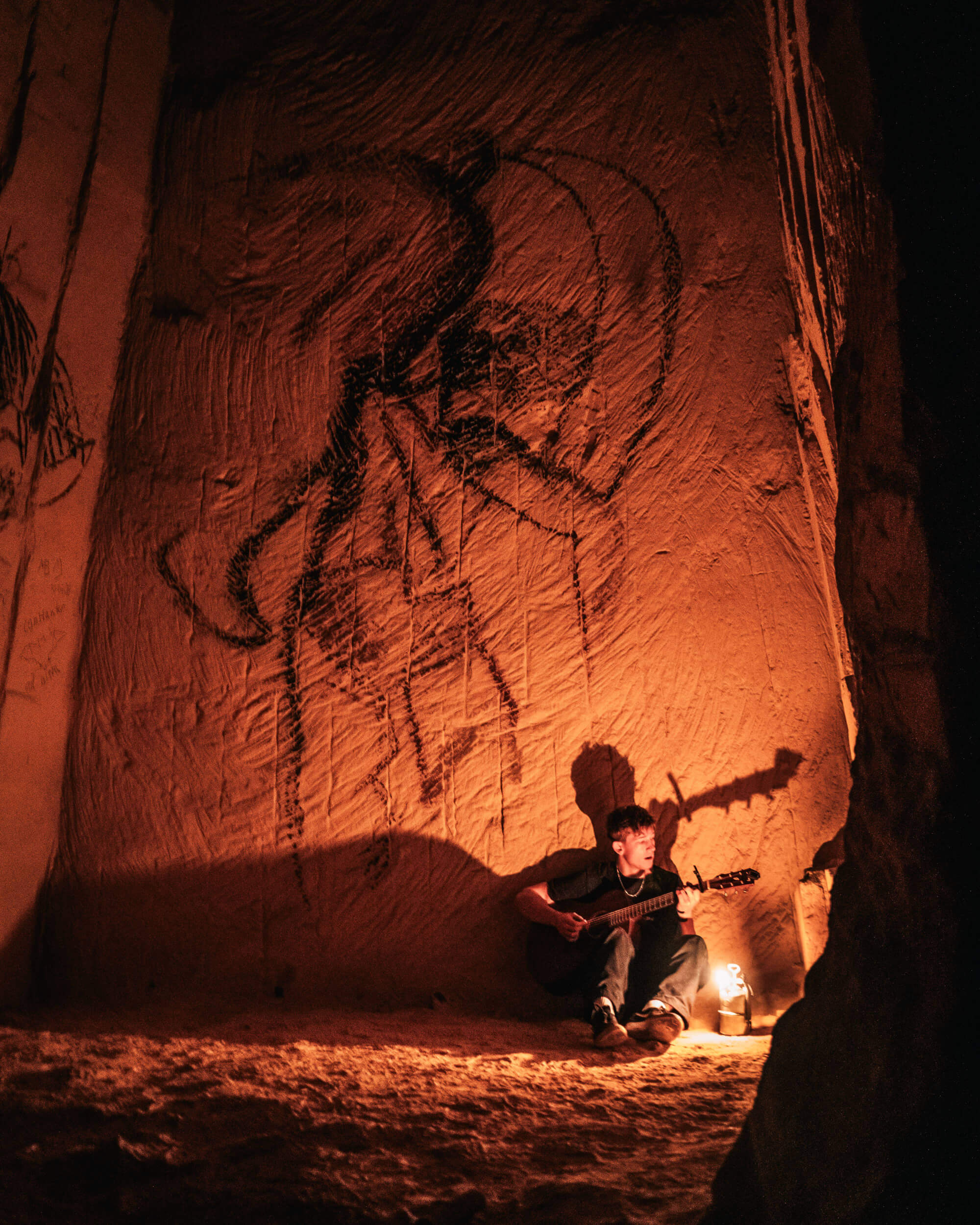 A young man sitting on the ground in a cave, playing an acoustic guitar next to a lantern, with ancient drawings on the cave wall behind him.
