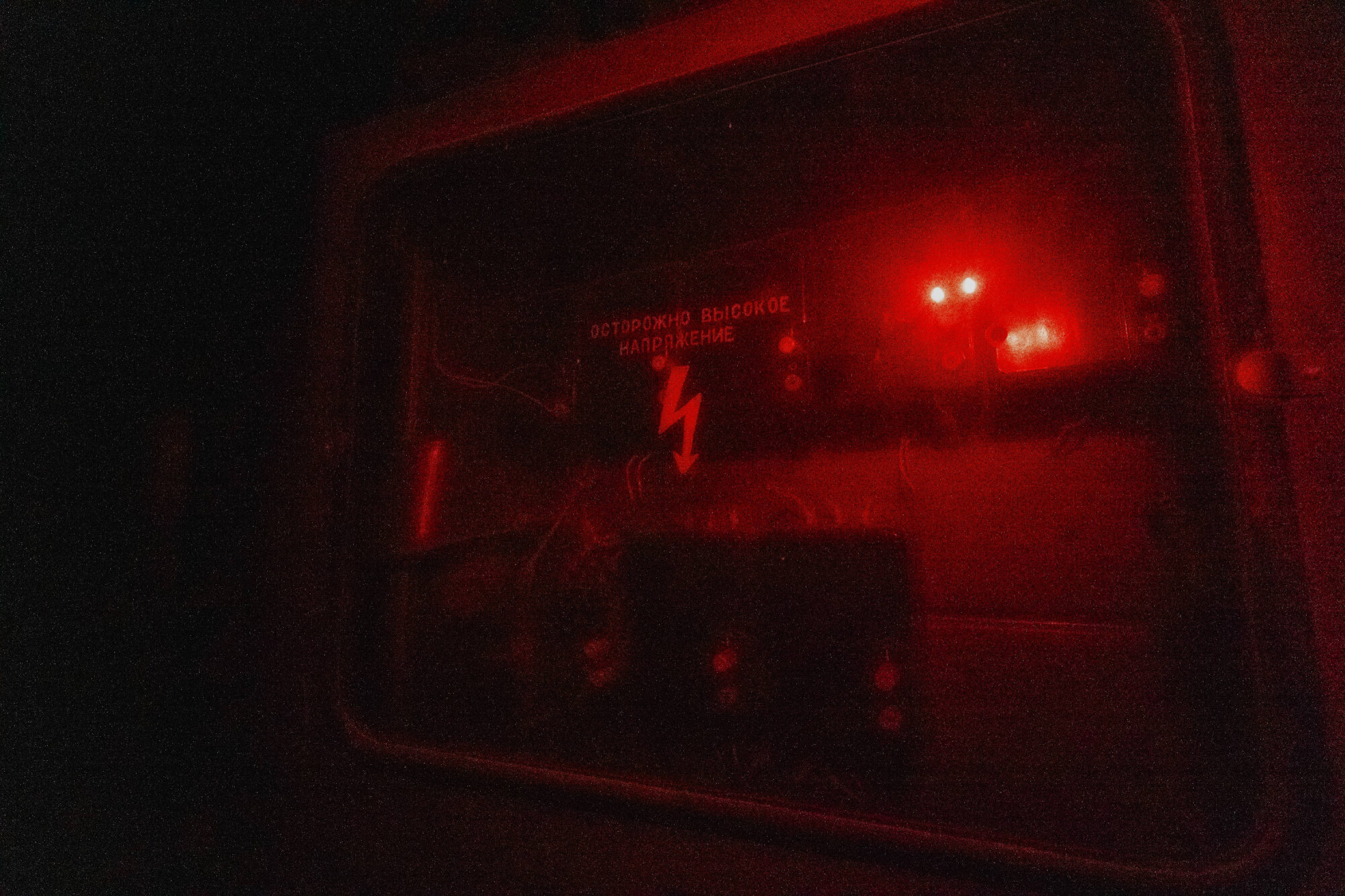 A dark control panel with a red glow, displaying a warning sign in Russian about high voltage, featuring illuminated lights and visible electrical components.