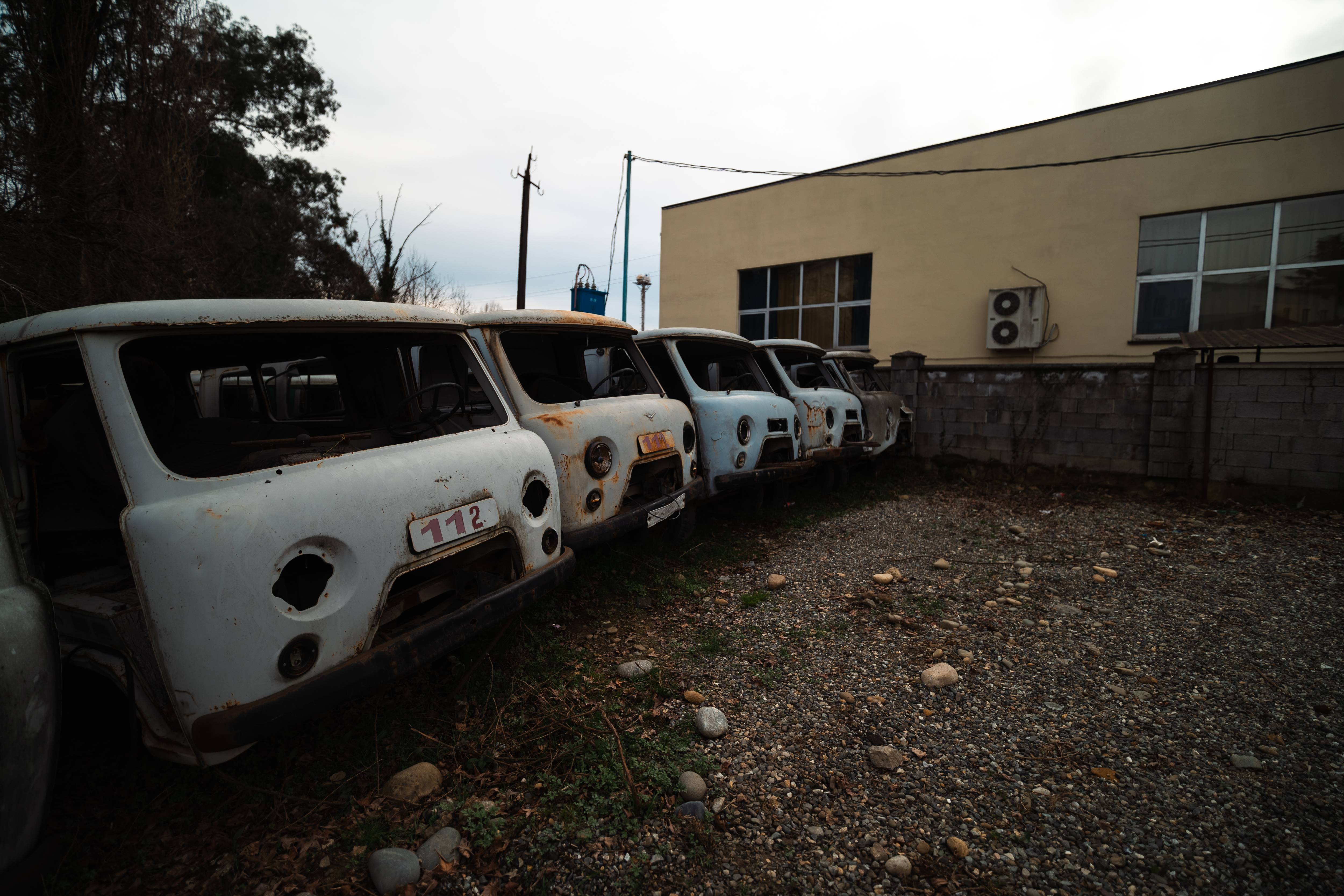 A row of abandoned, rusting vintage vehicles with missing windows, lined up in a gravel area next to a simple beige building. The sky is overcast, creating a gloomy atmosphere.