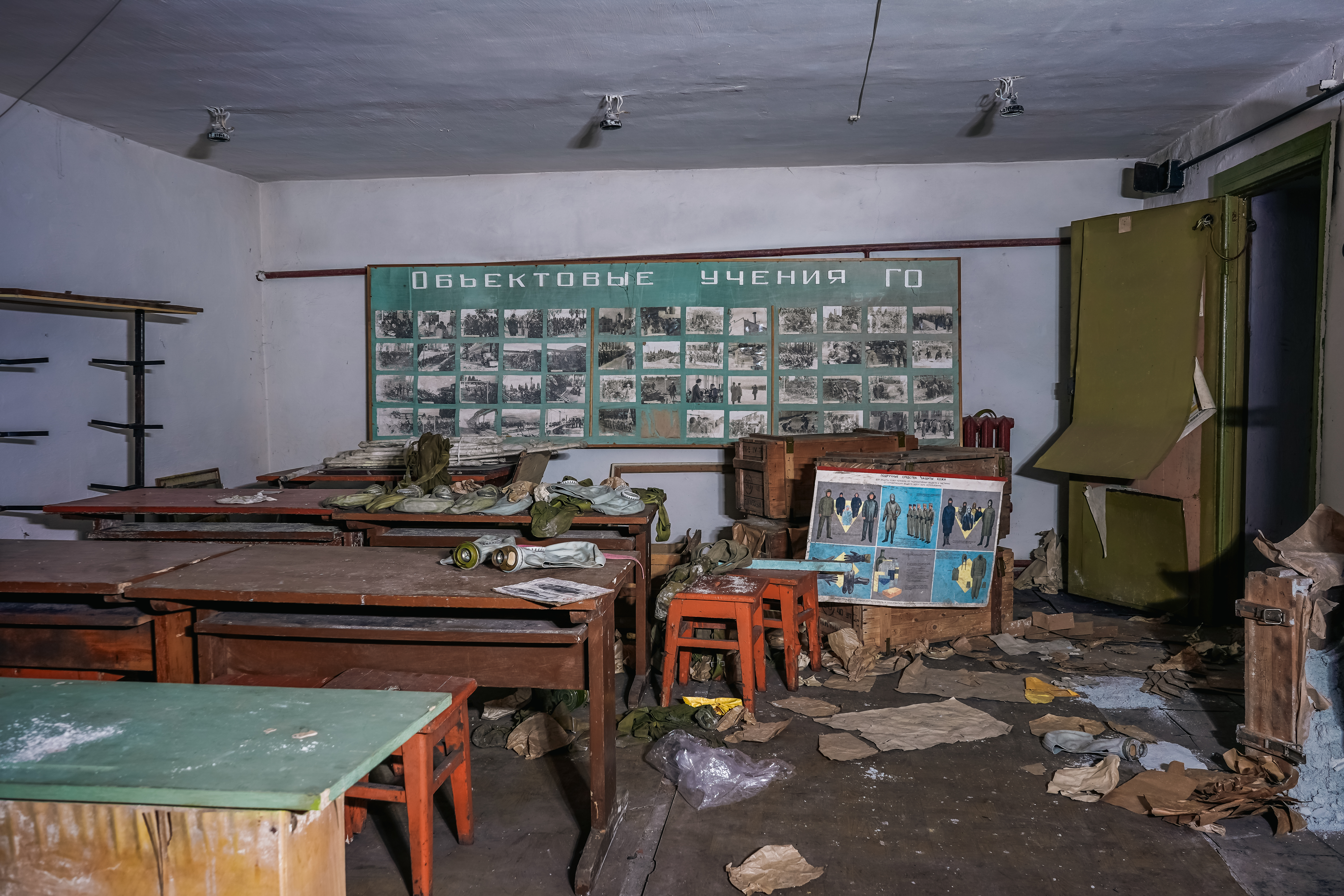 Interior of an abandoned classroom with dirty tables, scattered papers, and a green wall board covered with old photographs. The room is dimly lit, suggesting neglect.