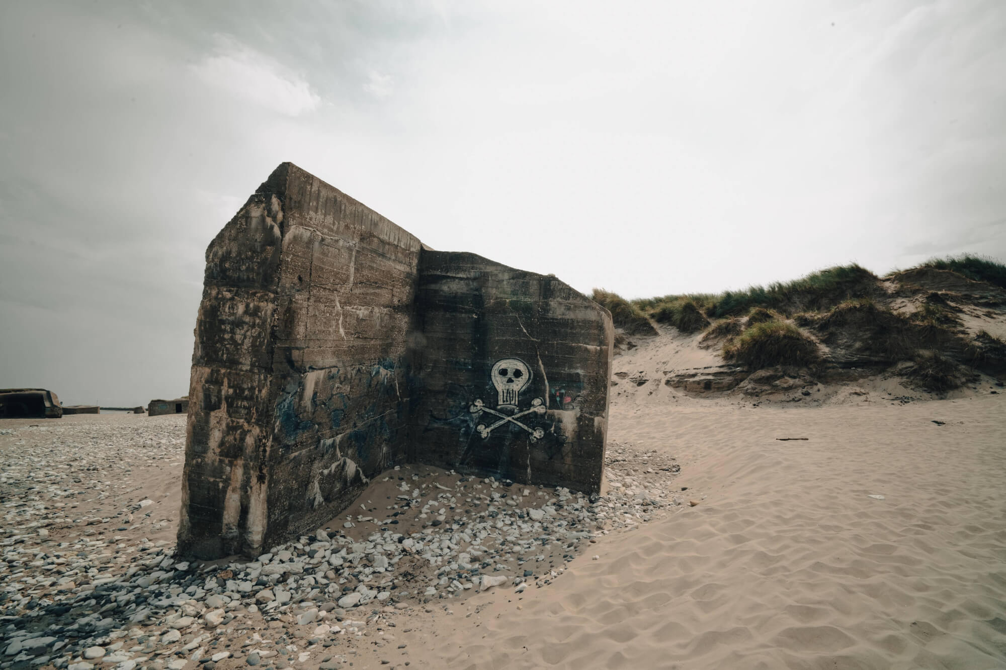 A weathered concrete structure with a pirate skull and crossbones graffiti stands on a sandy beach, surrounded by small pebbles, under an overcast sky with grassy dunes in the background.