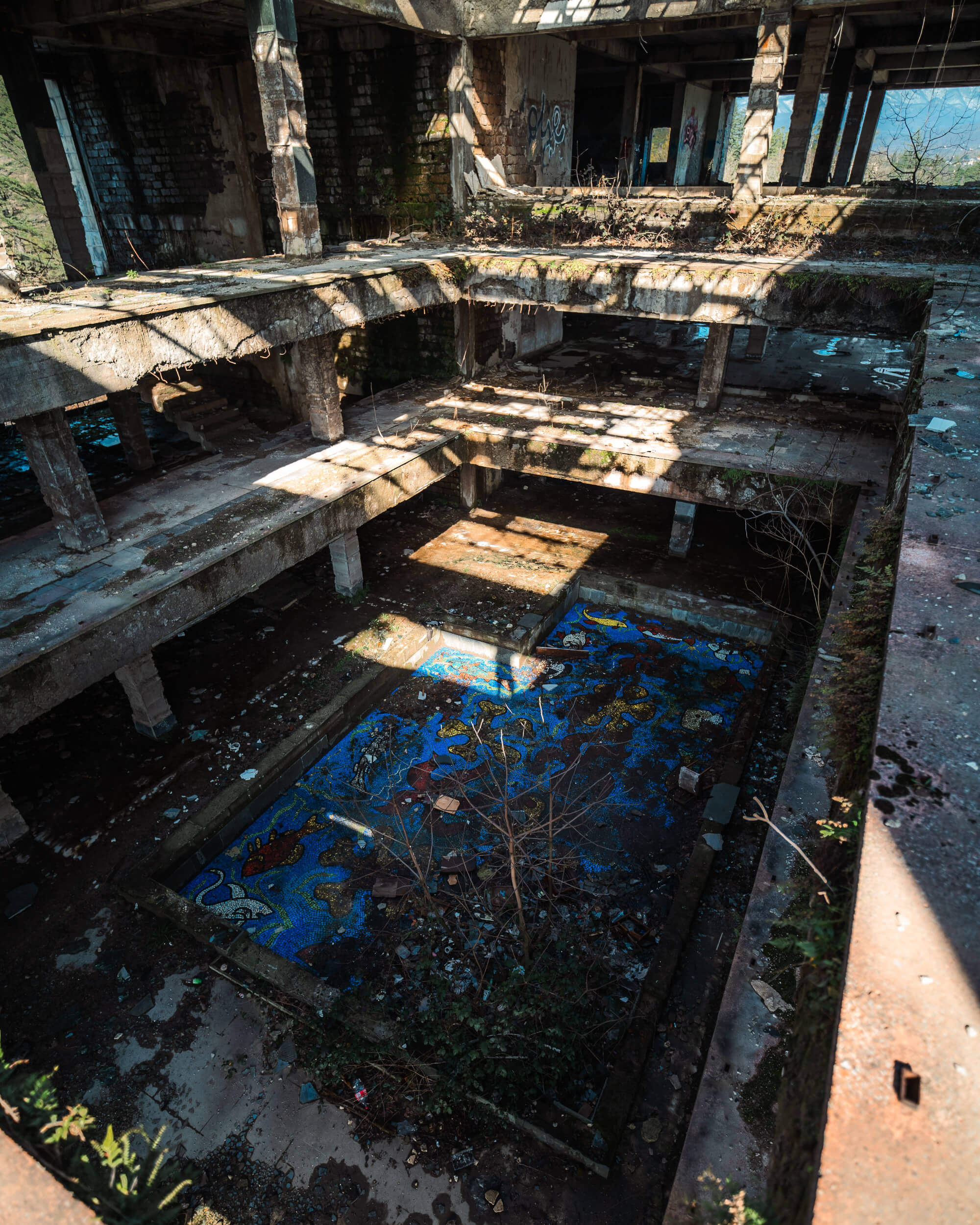 An overhead view of an abandoned building with broken floors and walls. Sunlight streams through the openings, highlighting a colorful mosaic in the center, surrounded by debris and overgrown plants.