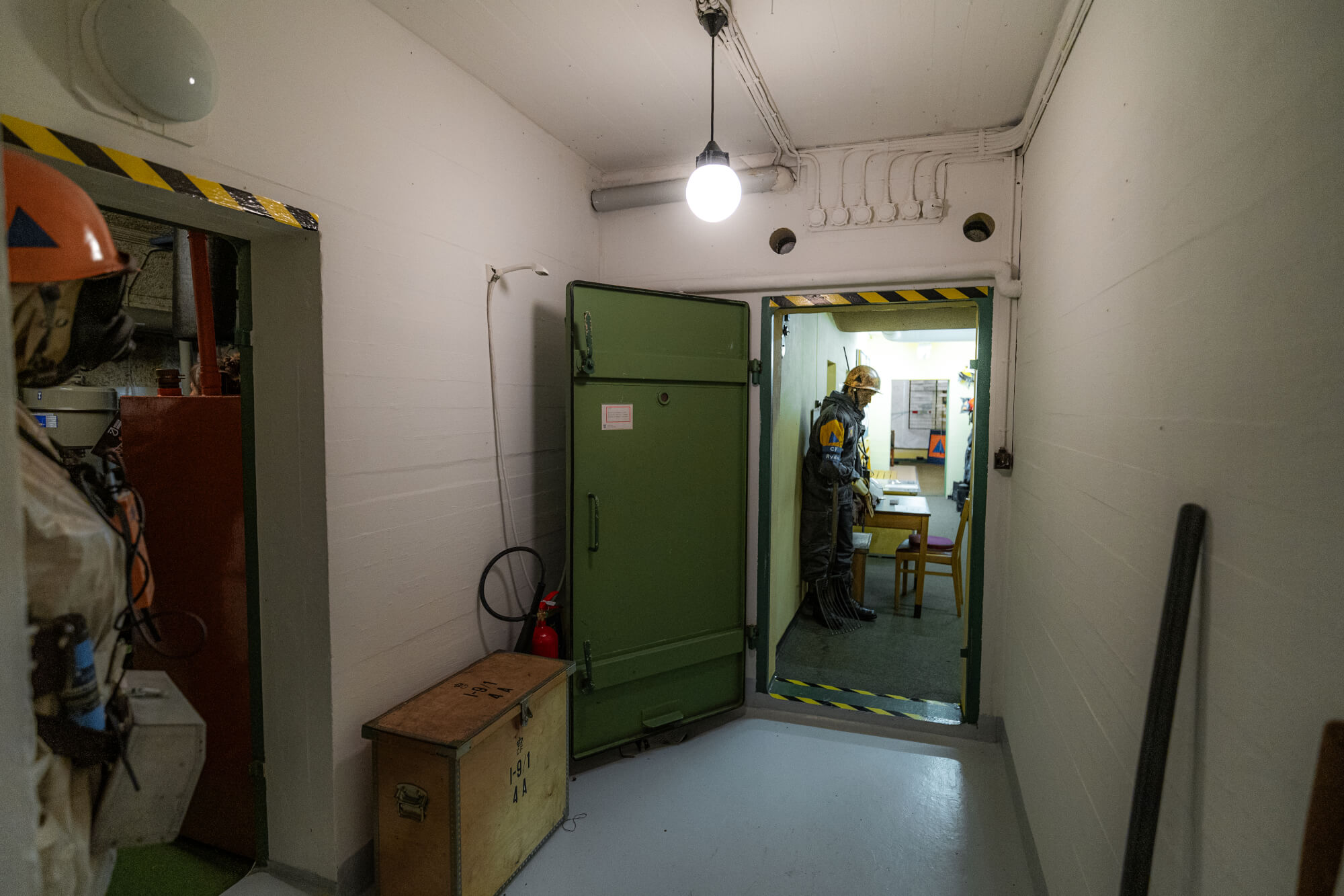 A dim corridor with a green door and a figure in protective gear standing inside a room. There is a wooden box on the floor, a fire extinguisher nearby, and the walls are lined with pipes and wiring. The scene evokes a bunker-like atmosphere.
