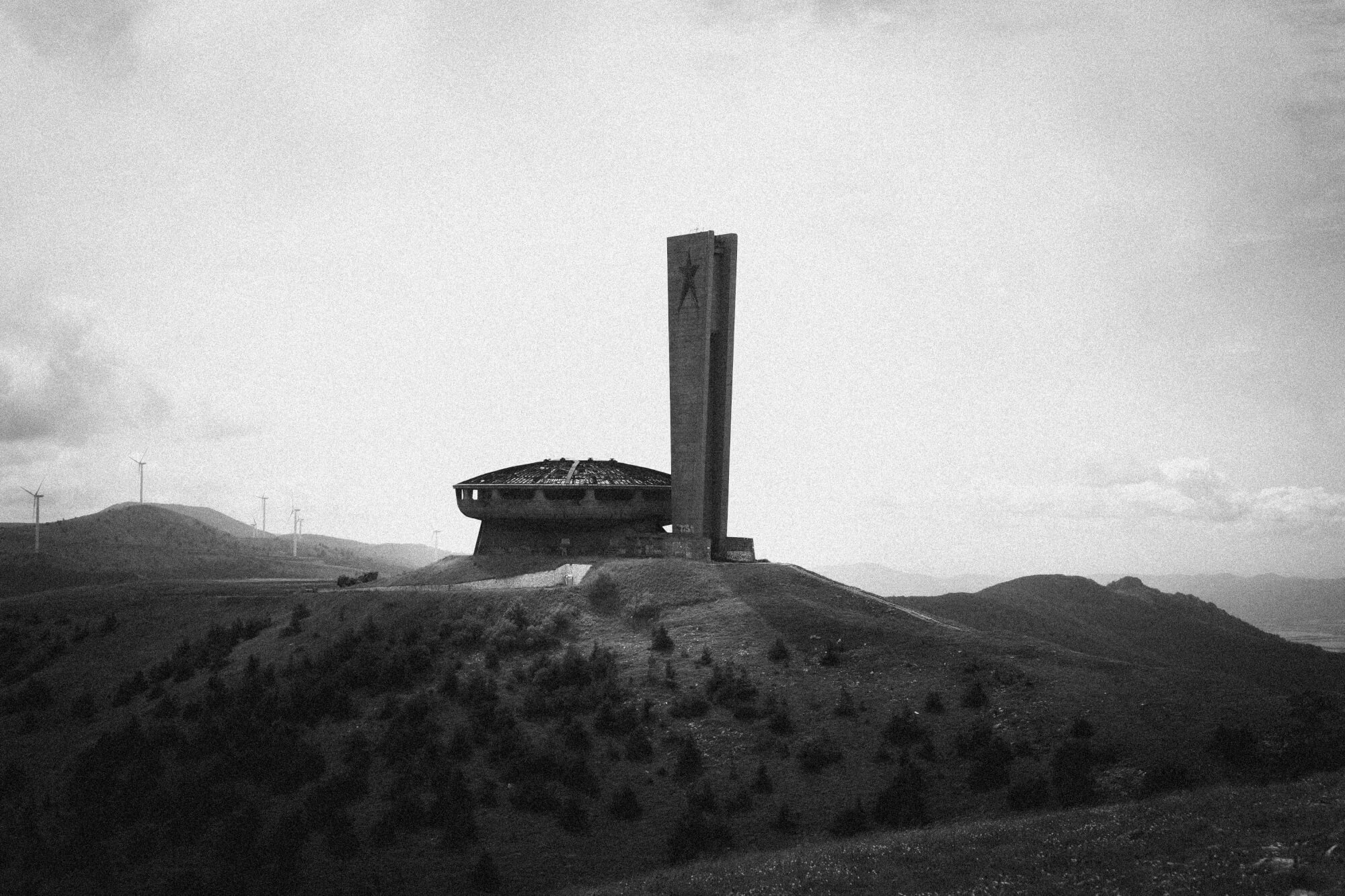 A desolate hillside with a circular, ruined building and a tall rectangular monument. Wind turbines are visible in the background against a cloudy sky.