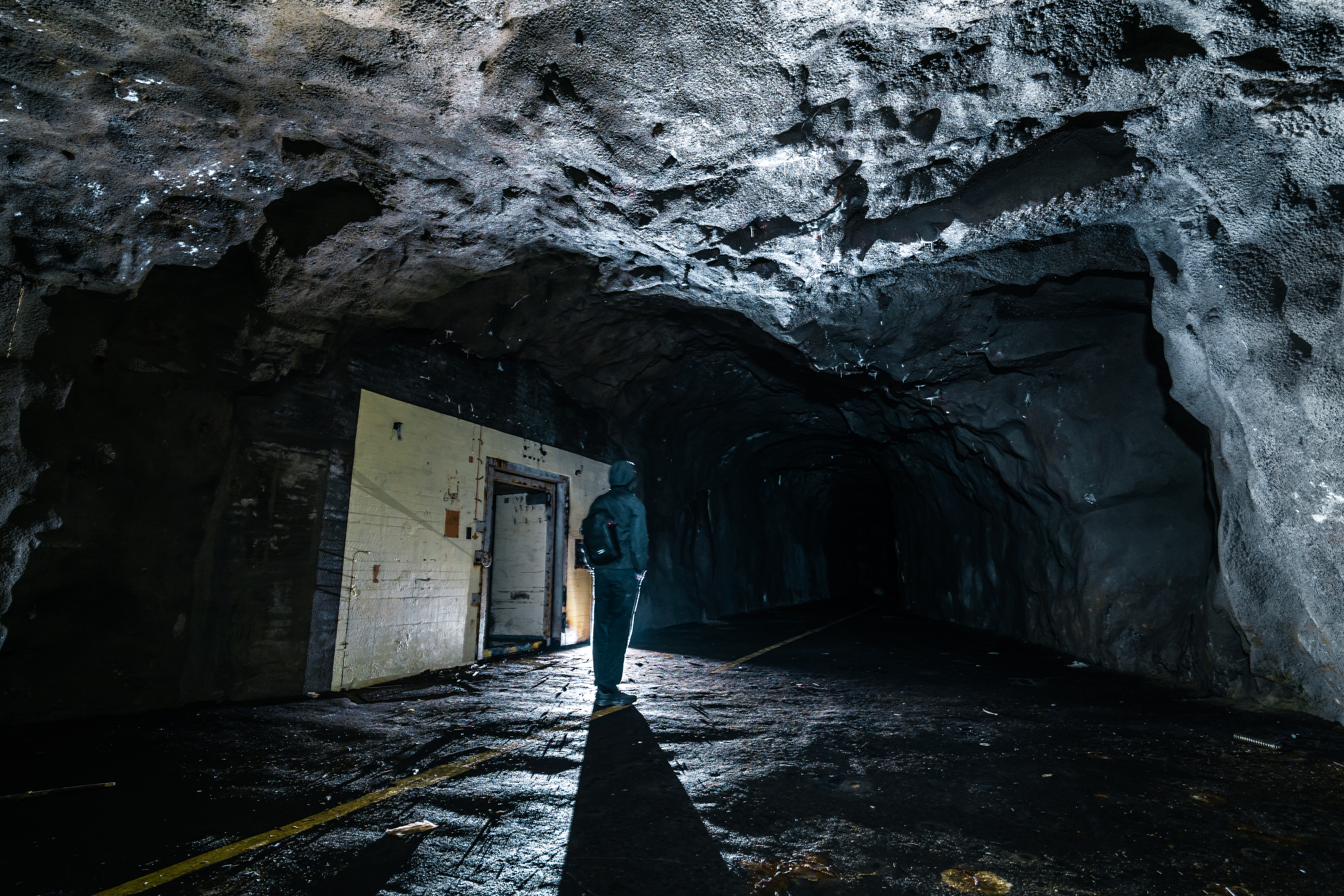 A dark tunnel with a person in a hooded jacket standing in the foreground, illuminated by overhead light. The rocky walls show moisture and texture, while a faint path runs through the center.