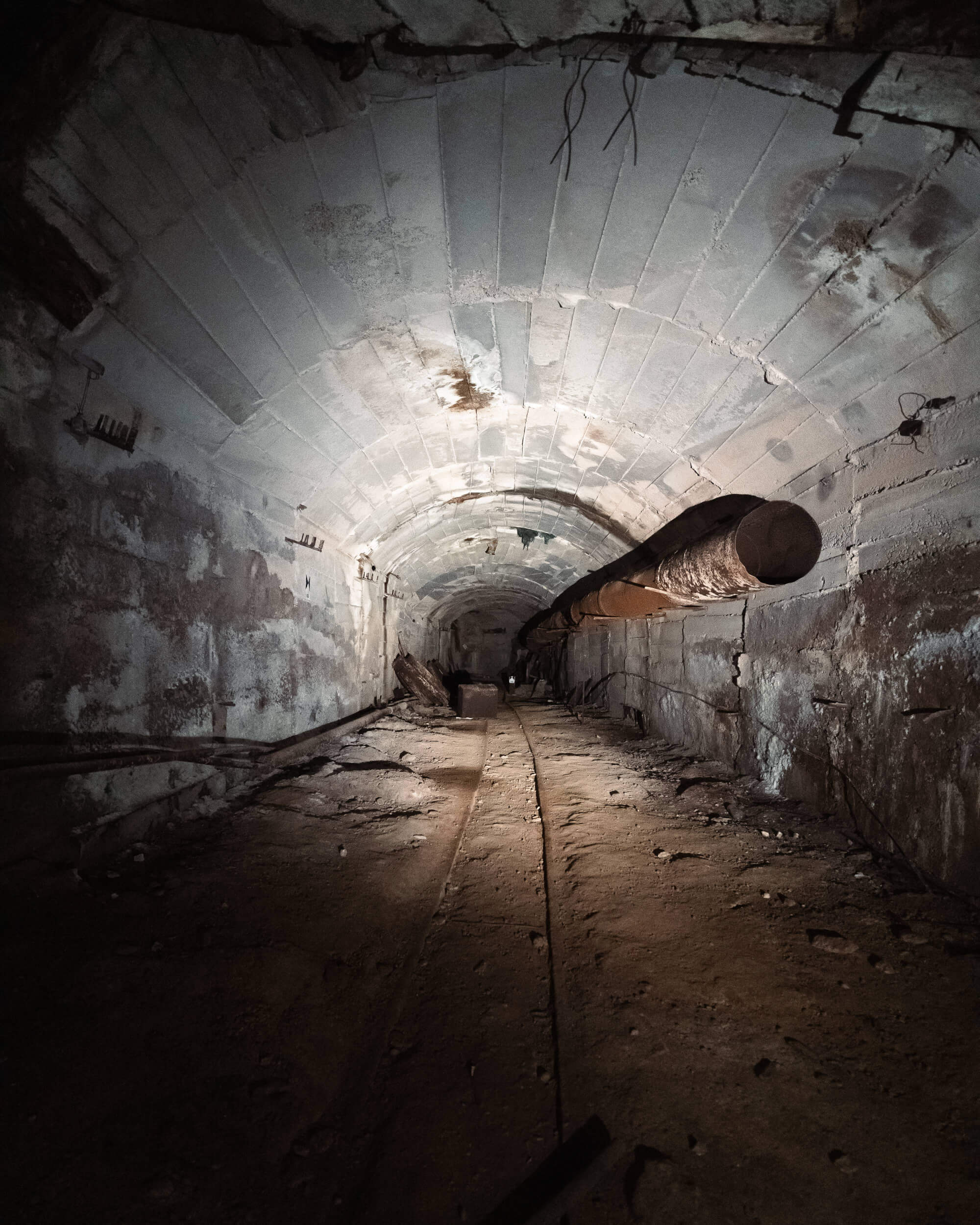 A dimly lit underground tunnel with an arched ceiling and rough walls. A rusted pipe runs along one side and railway tracks are visible on the ground, surrounded by dirt and debris.