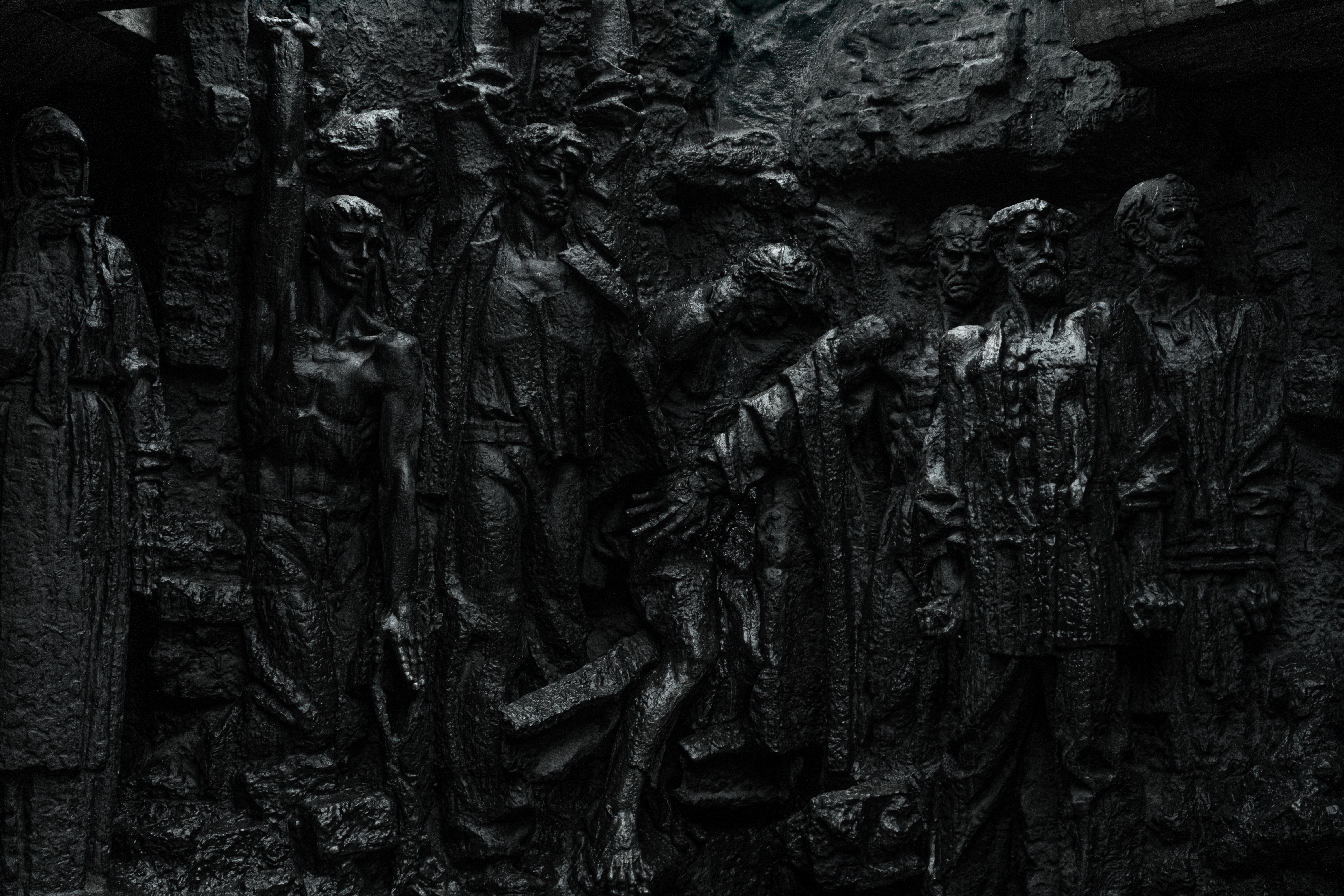 A dark, textured wall with sculpted figures depicting various human emotions and expressions, including strength and despair, against a somber background.