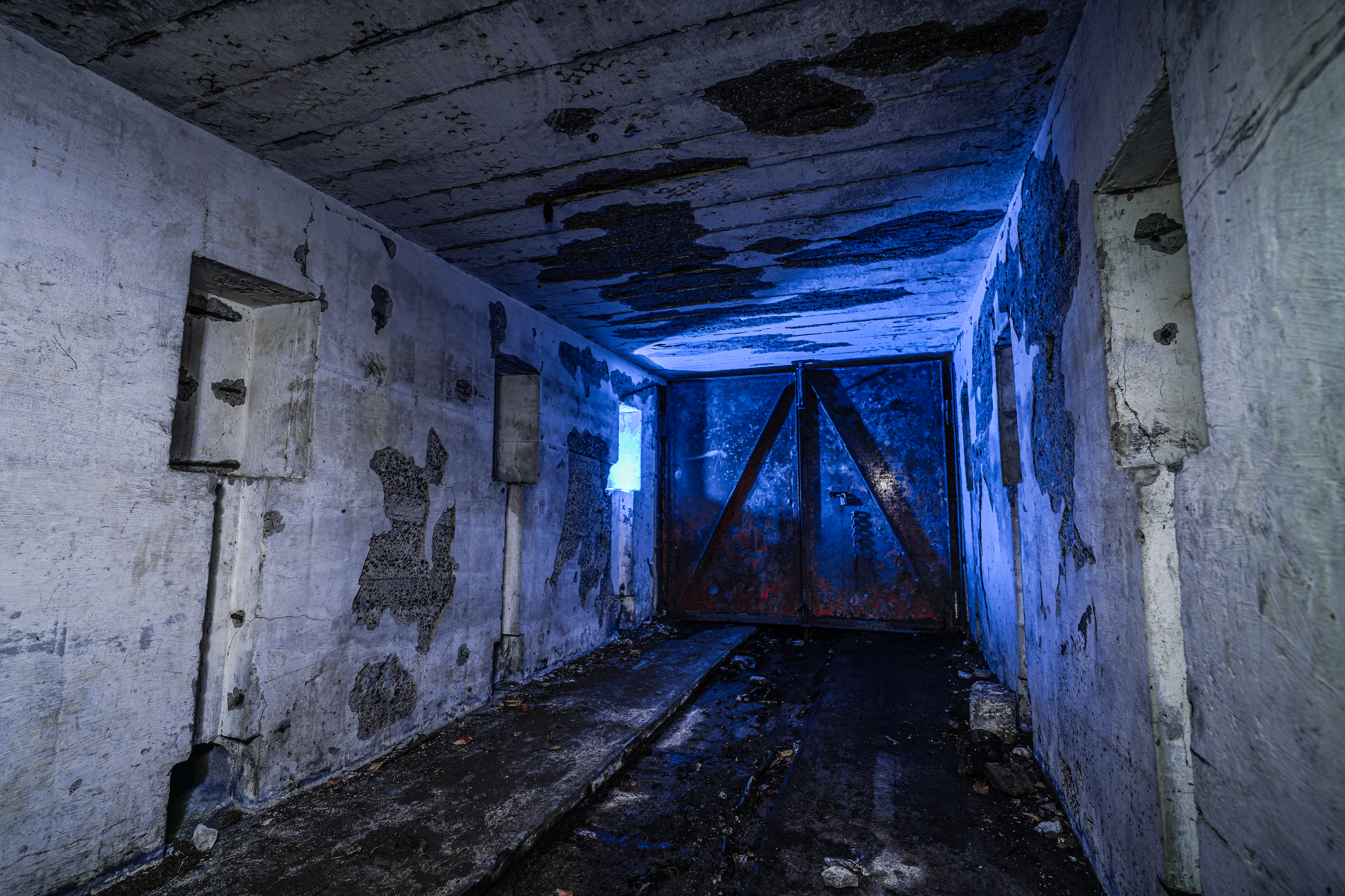 An abandoned, dimly lit room with gray concrete walls showing signs of decay. A rusted metal door is positioned at the end, and a small window emits a blue light, highlighting the textures of the surroundings.