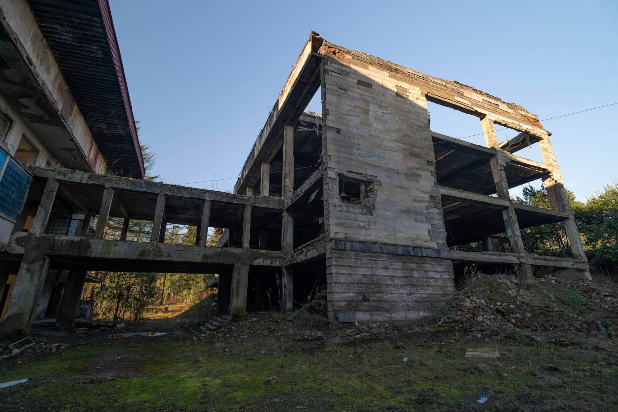 A partially collapsed concrete building with exposed floors and walls, surrounded by overgrown greenery and moss-covered ground. The sky is clear and blue above.