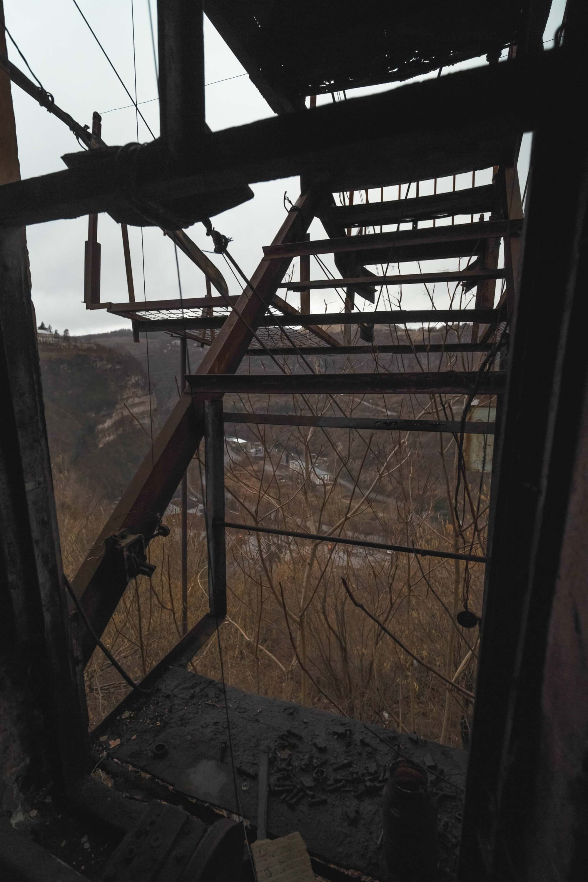 An interior view through a window of a dilapidated structure, featuring a rusted staircase leading outside surrounded by barren branches and a cloudy sky.