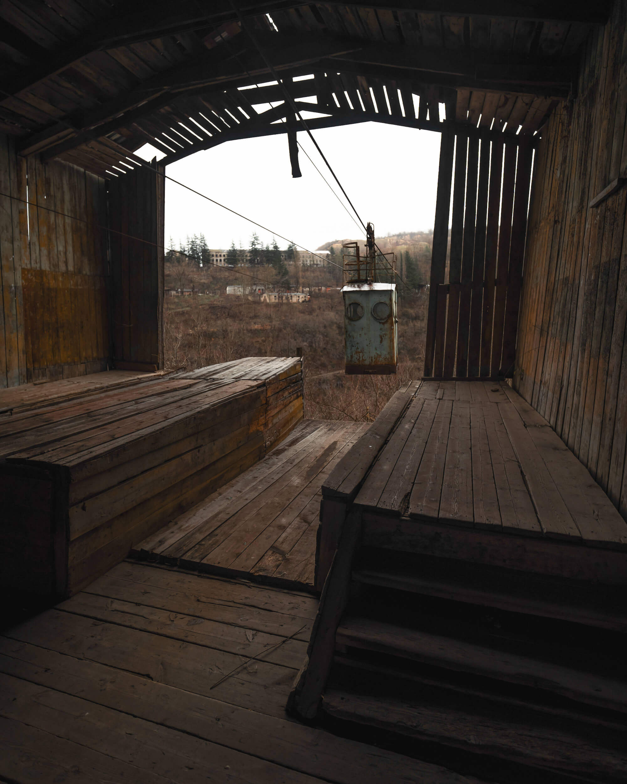 Interior view of an abandoned building with wooden floors and walls. A rusted cable car hangs suspended by cables, with soft light illuminating the area.
