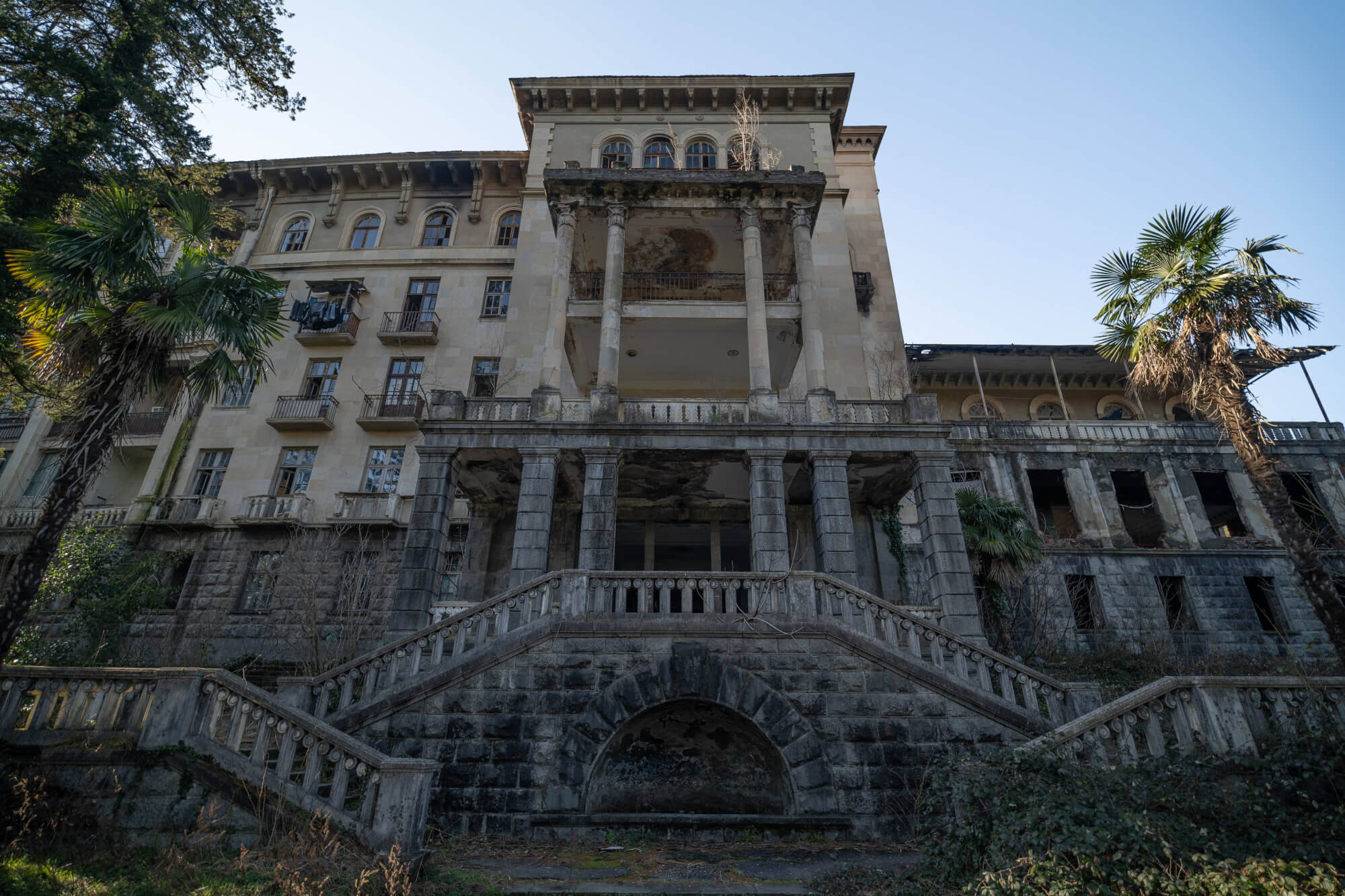 A dilapidated building with large columns and multiple balconies. Some windows are broken, and there's greenery on the ground and around the structure. The sky is clear and blue.