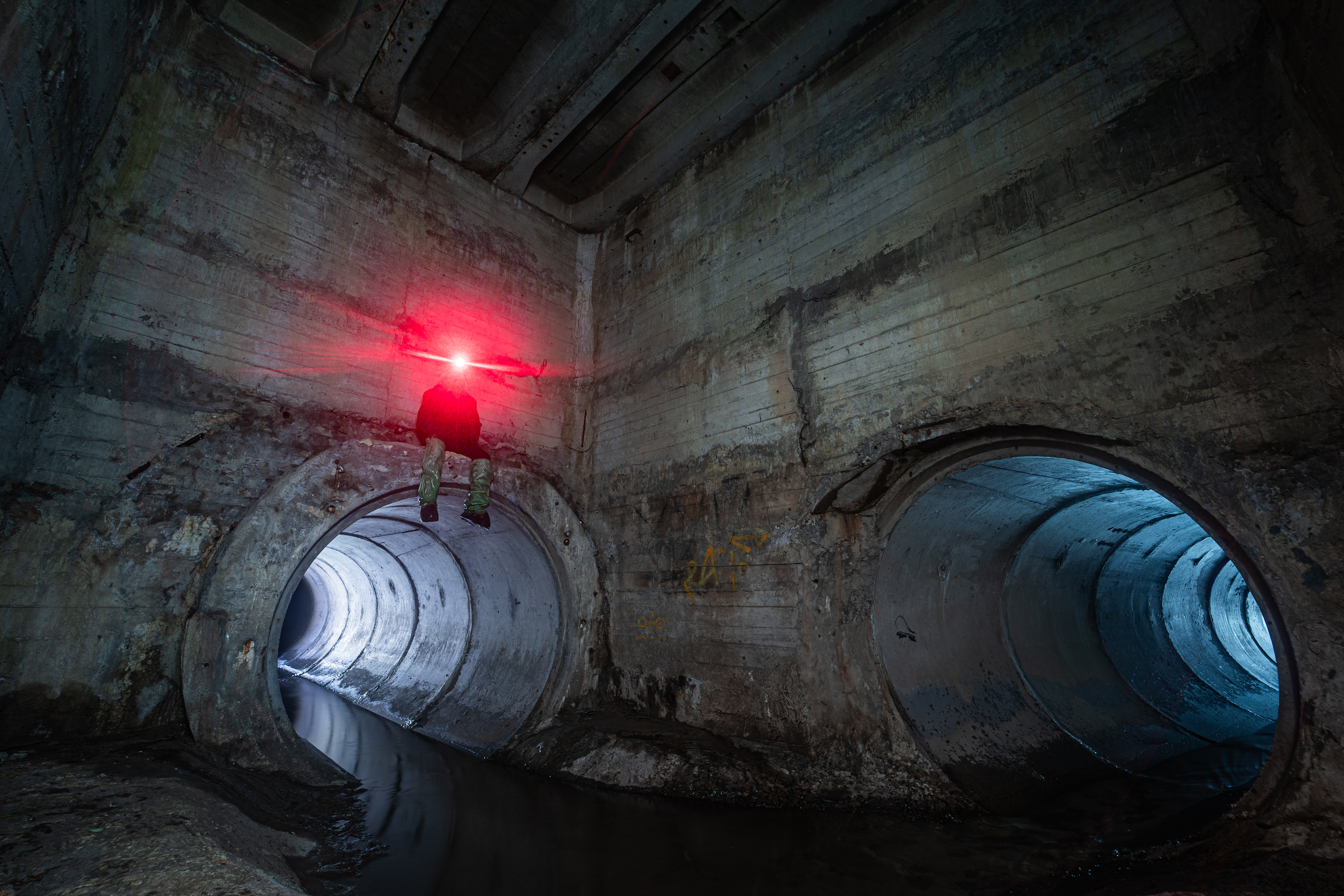 An underground tunnel with two large circular openings, one of which is illuminated in blue. A person wearing dark clothing and a bright red light on their head is sitting on the edge of the wall near the tunnel entrance.