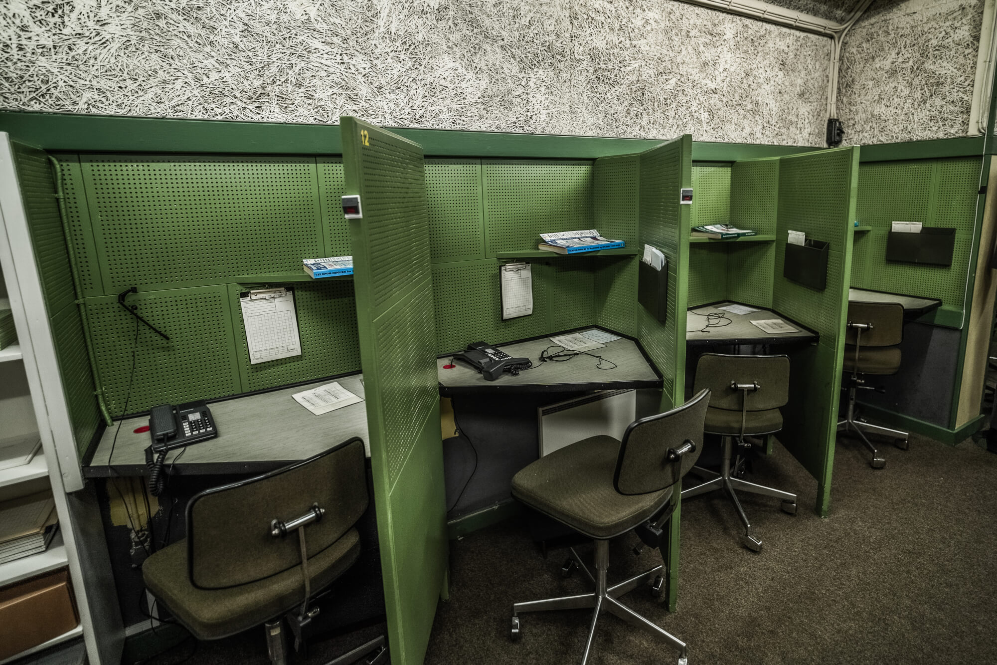 A series of green cubicles with simple desks featuring telephones and papers. The walls above are patterned, and the floor is carpeted, creating a subdued workspace environment.