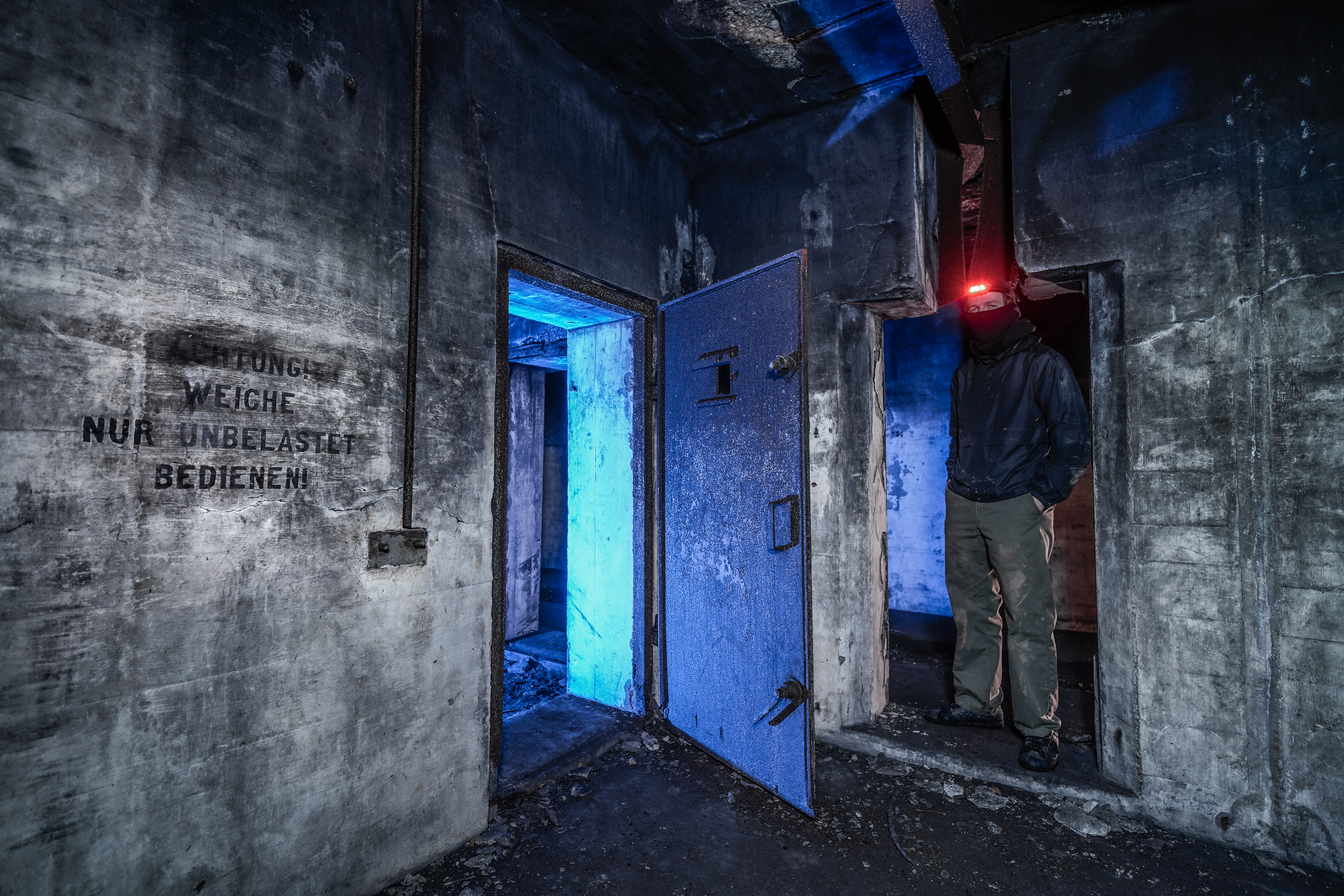An abandoned industrial interior with a man standing in front of two doorways; one doorway is lit with blue light, while the other is dark. The walls are dirty and have graffiti. The man is wearing dark clothing and a headlamp.