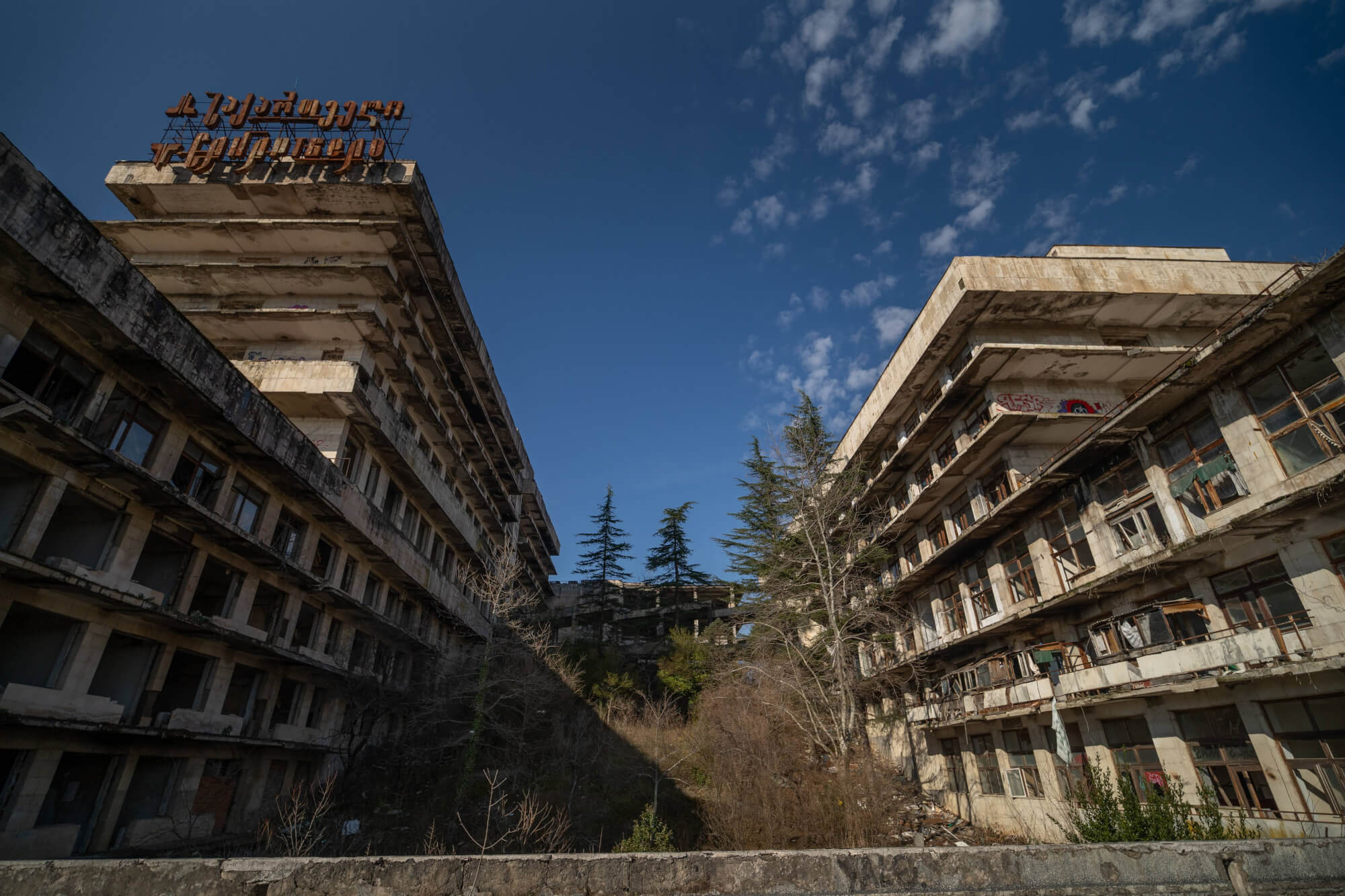 A pair of abandoned buildings with broken windows and a weathered facade, surrounded by sparse trees and overgrown vegetation under a bright blue sky.